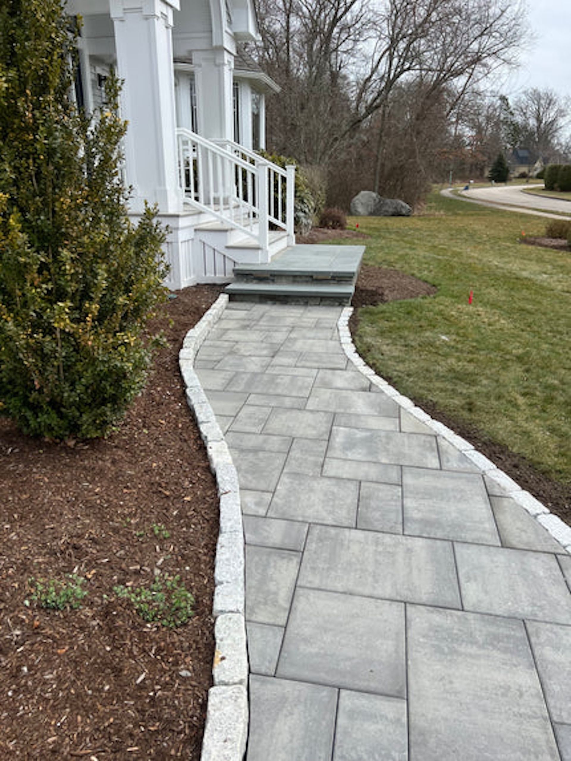 A stone paver walkway with granite edging leading up to the white steps and porch of a house.