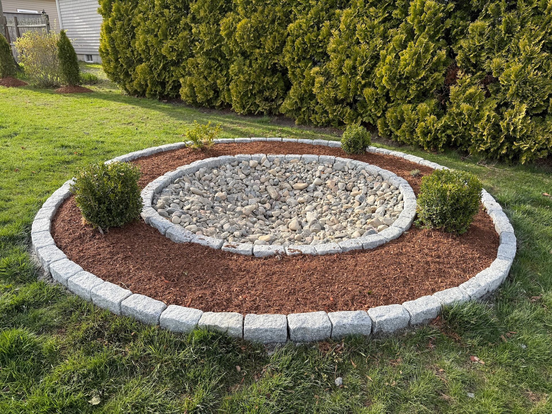 A circular garden bed featuring an inner ring of river rocks, a mulch border, and stone edging, set in a grassy yard.