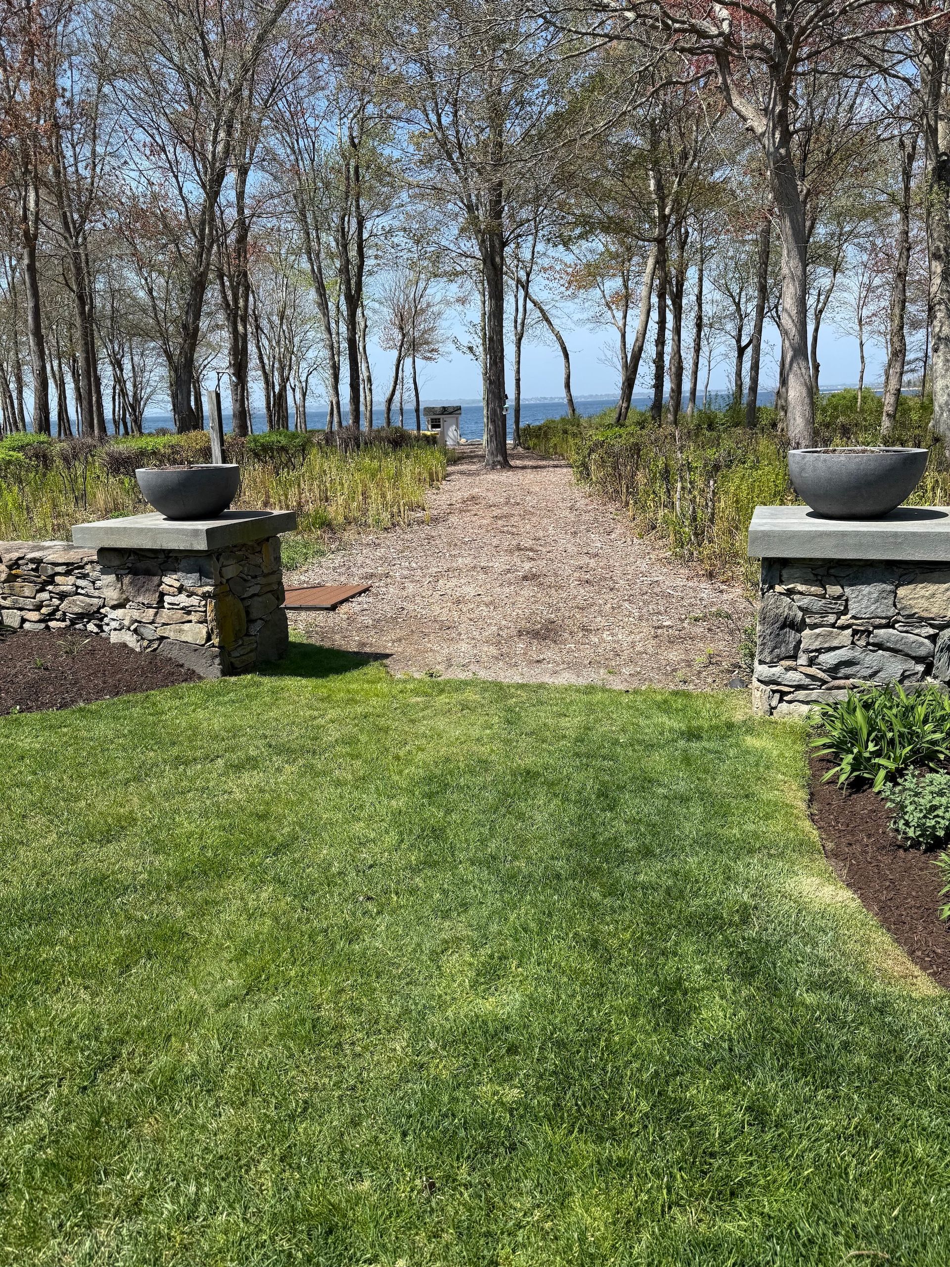 A stone-paved path flanked by two stone pillars with planters leads through trees toward a distant view of the ocean.