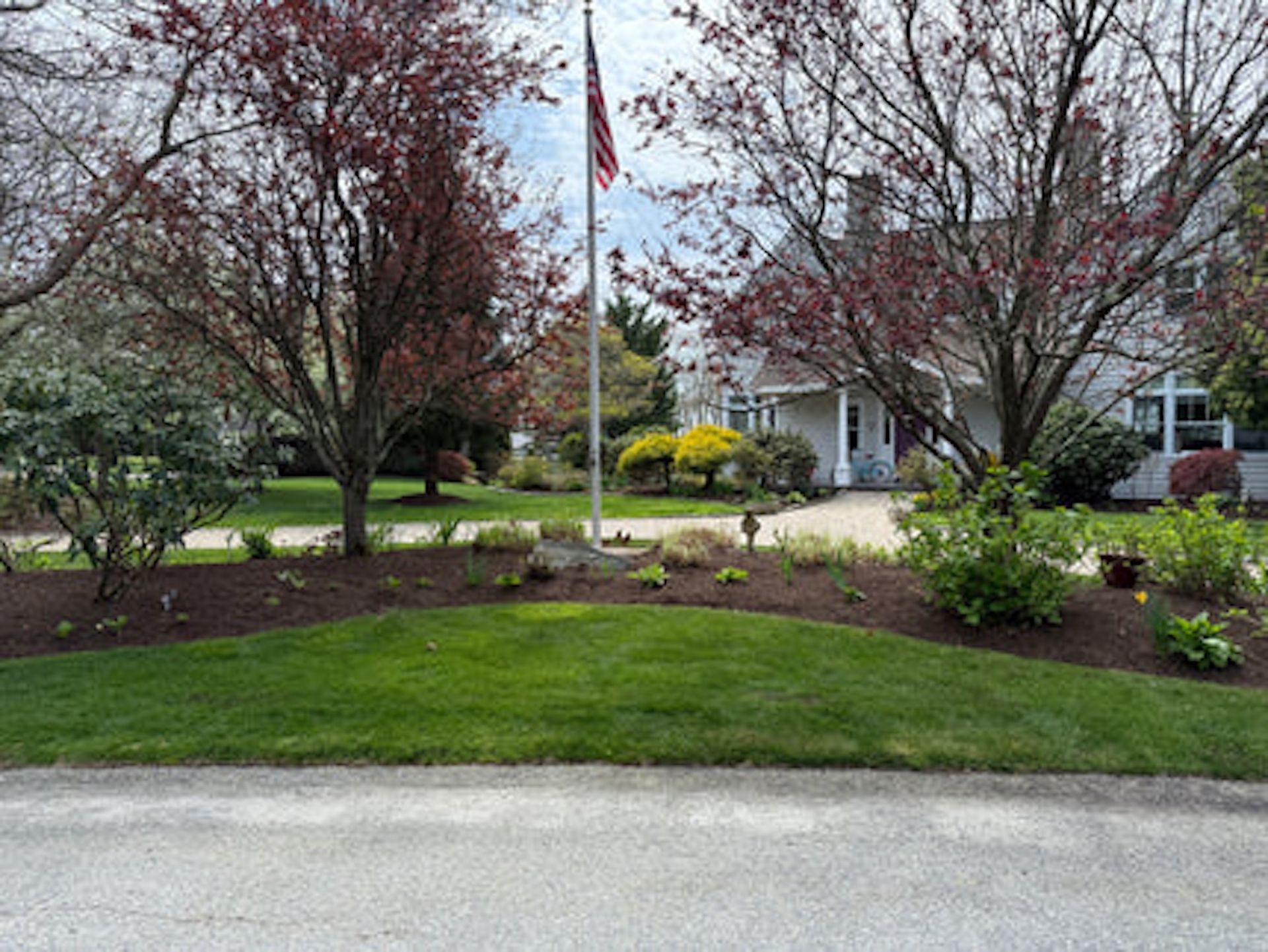 A flag flies in front of a house, surrounded by trees and a landscaped garden bed with mulch and a green lawn.