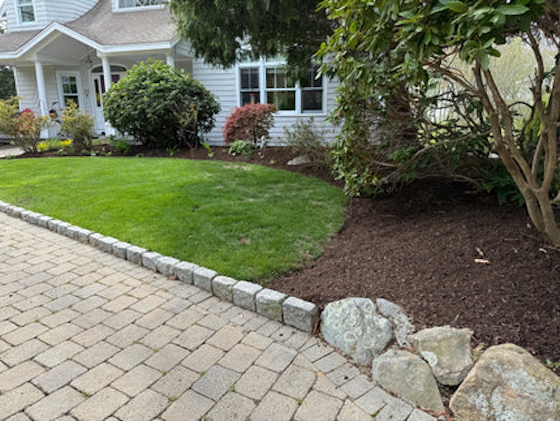 A suburban home front with a paved walkway, a manicured green lawn, dark mulch flower beds, and large landscape stones.