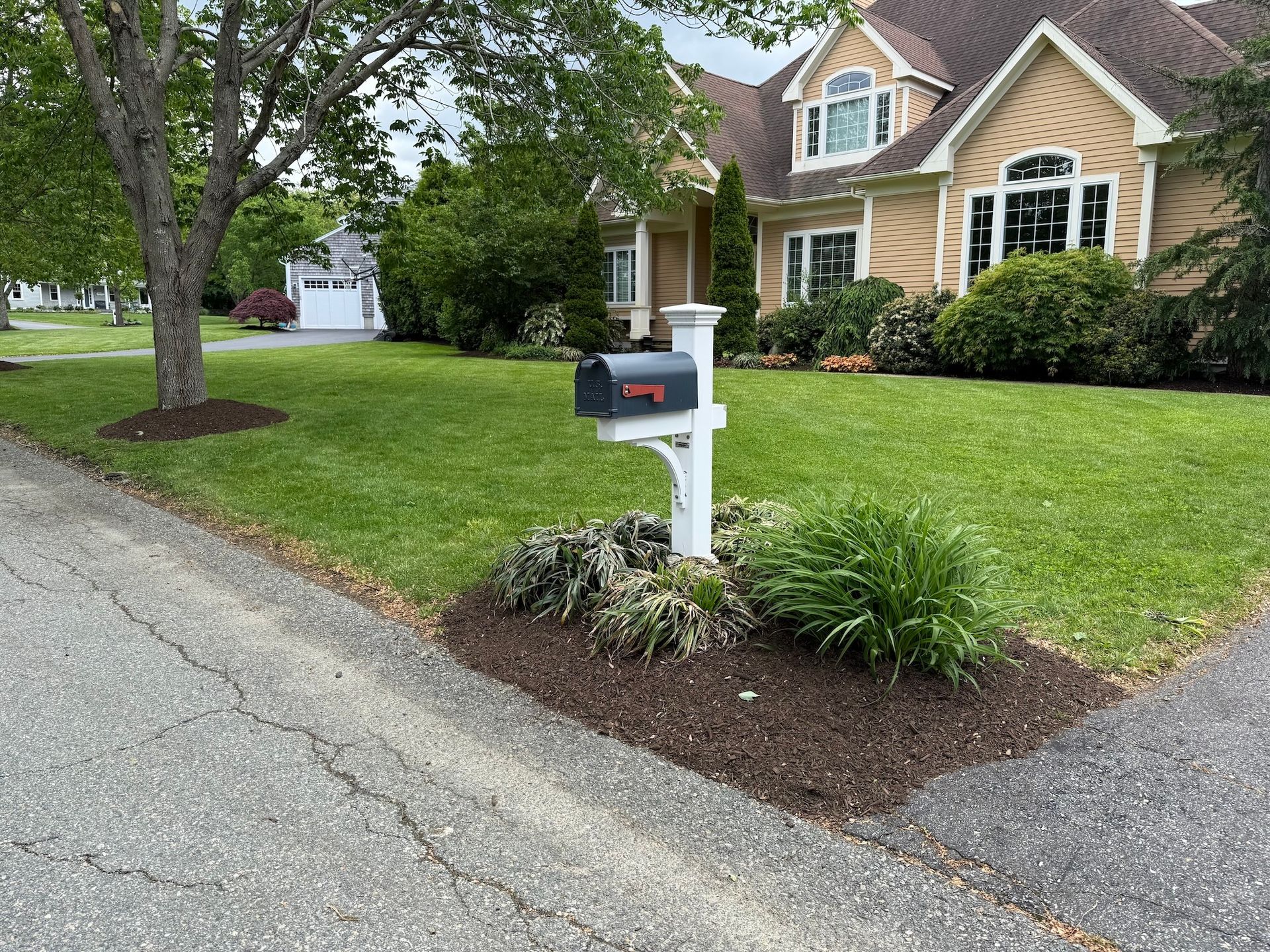 A curbside mailbox with a dark box on a white post, situated in a mulched garden bed in front of a tan suburban home.