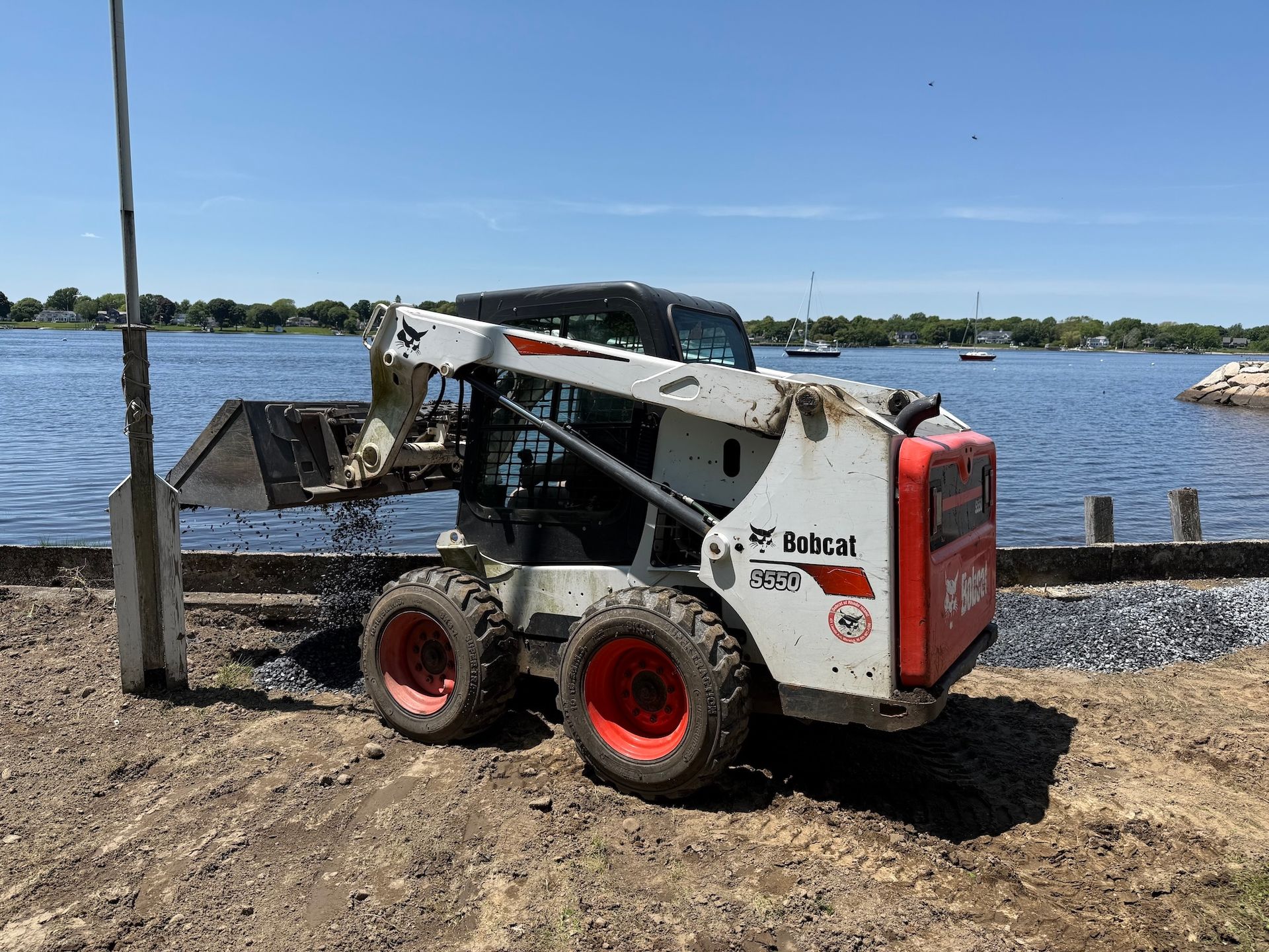 A white Bobcat skid-steer loader operating with its bucket lowered near a shoreline beside a body of water.