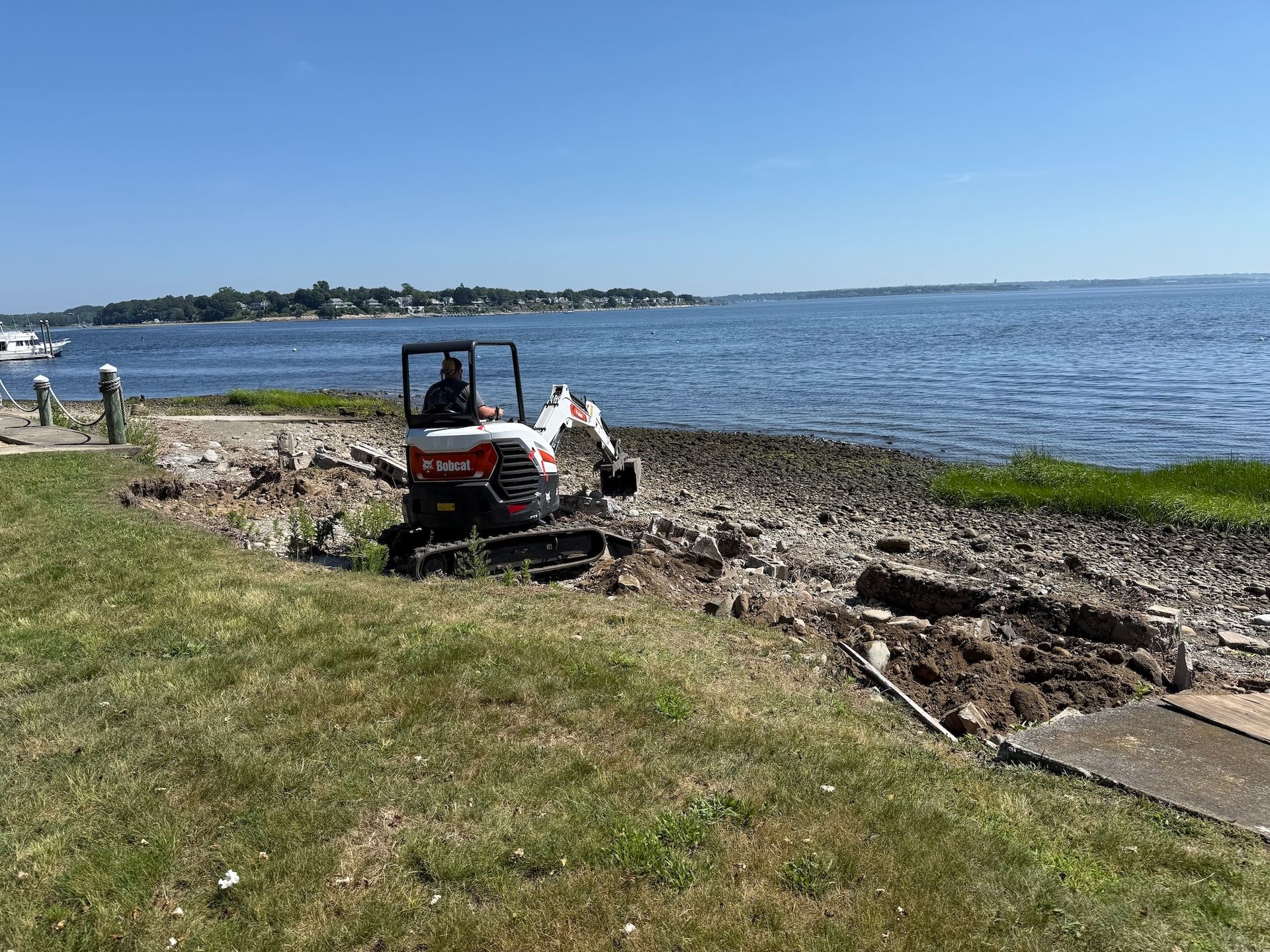 A white Bobcat mini-excavator works on a rocky shoreline next to a large body of water on a sunny day.