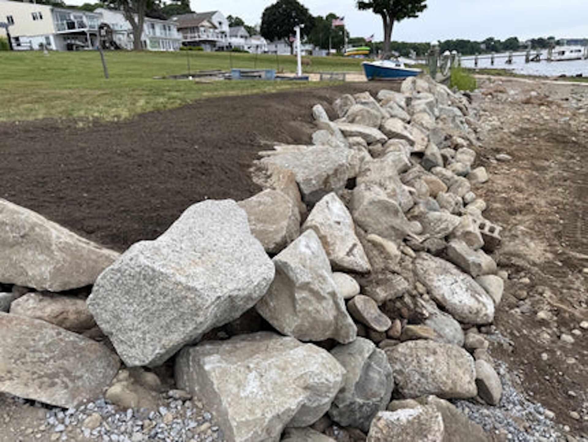 A newly constructed stone seawall separates a leveled dirt lawn from a sandy shoreline and water.