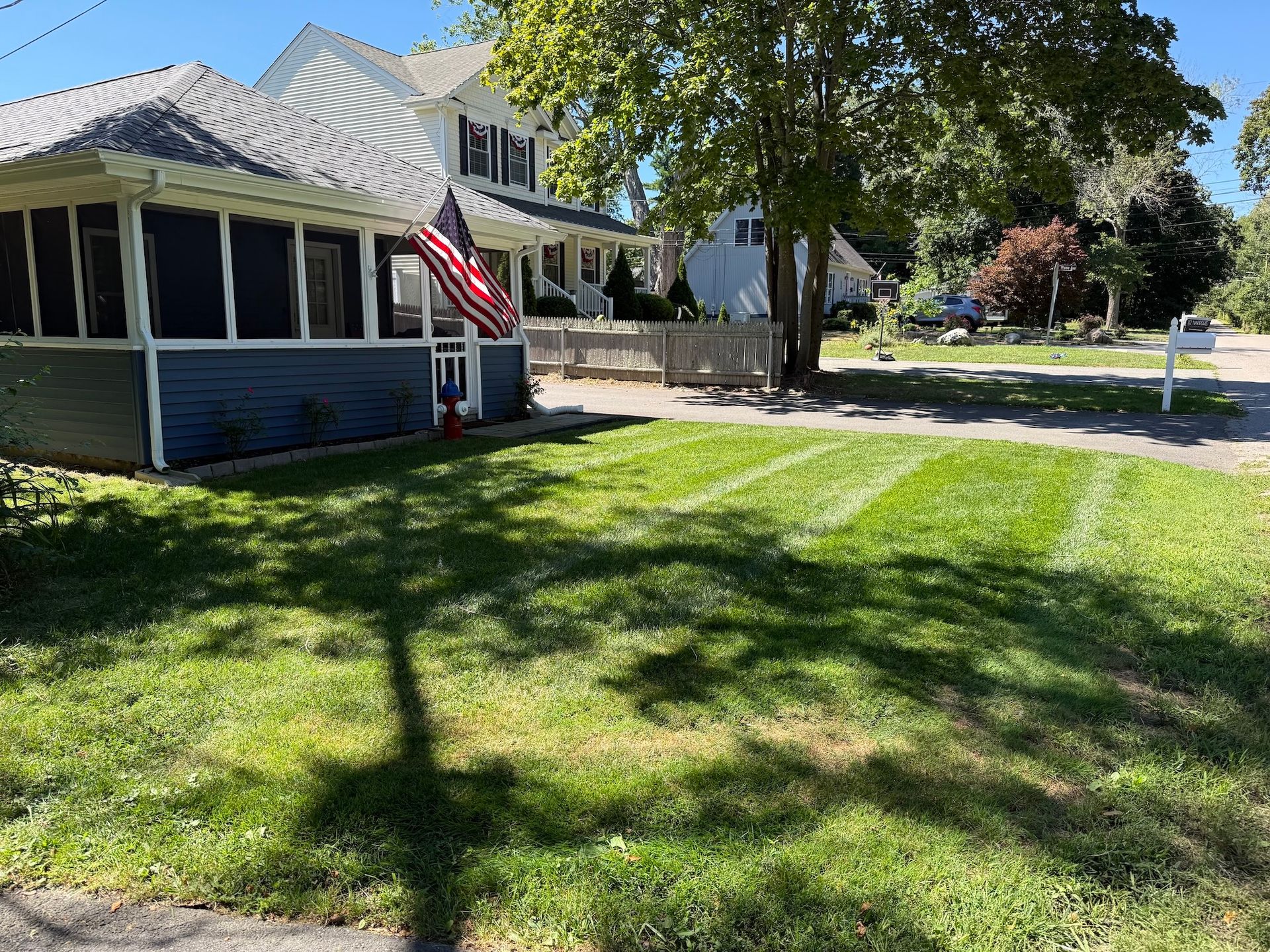A blue house with a white-trimmed porch and an American flag stands next to a sunny front lawn with visible mower stripes.