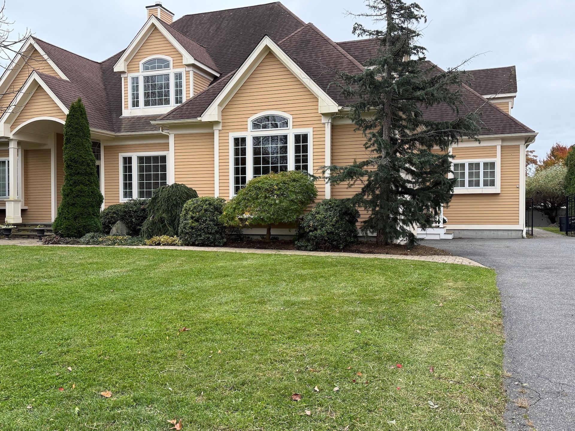 A large, tan suburban house with a dark brown roof, multiple gables, and a gravel driveway in front of a green lawn.