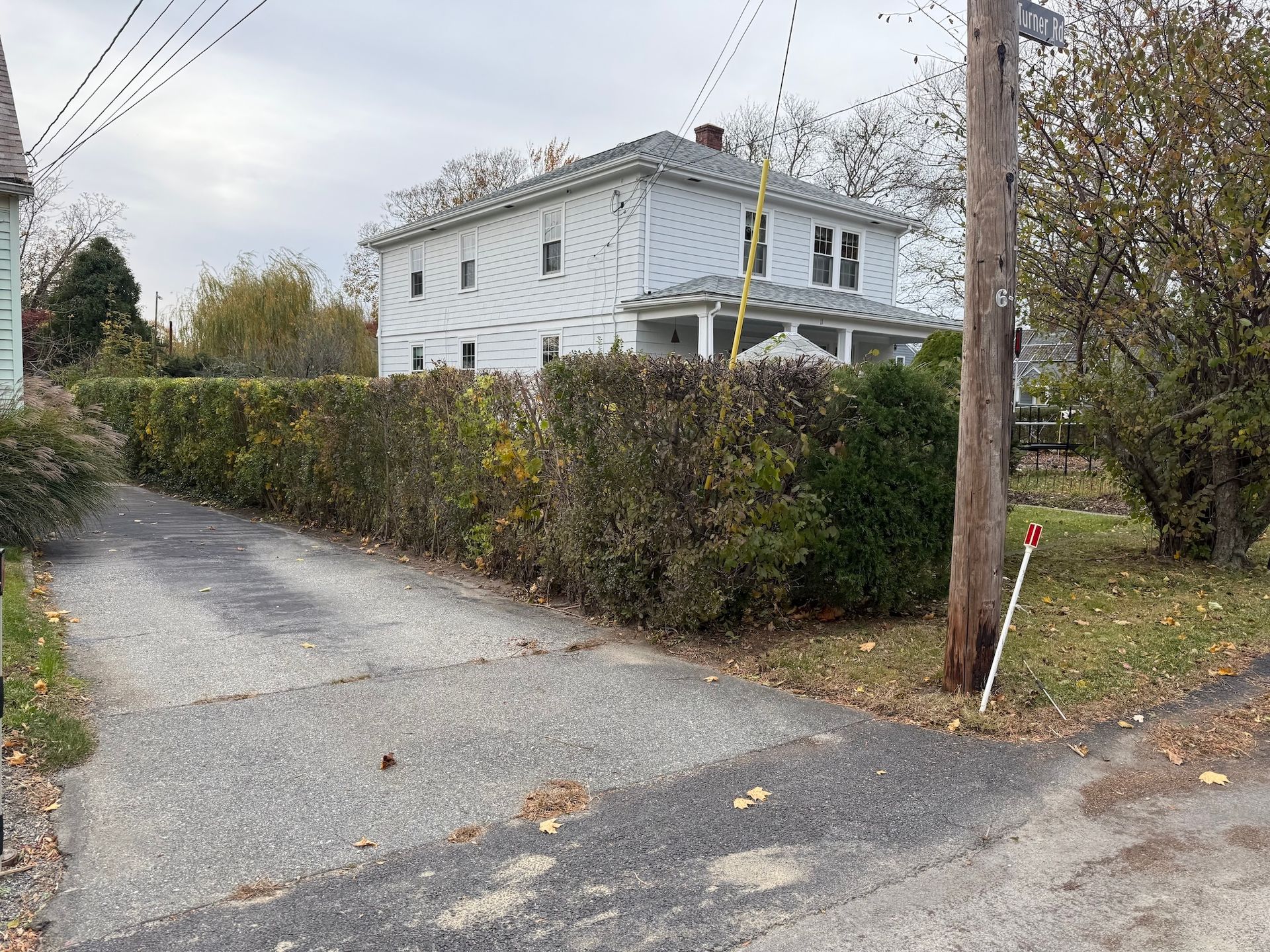 A two-story white house viewed from a gravel driveway, partially obscured by a tall, dense hedge under a cloudy sky.