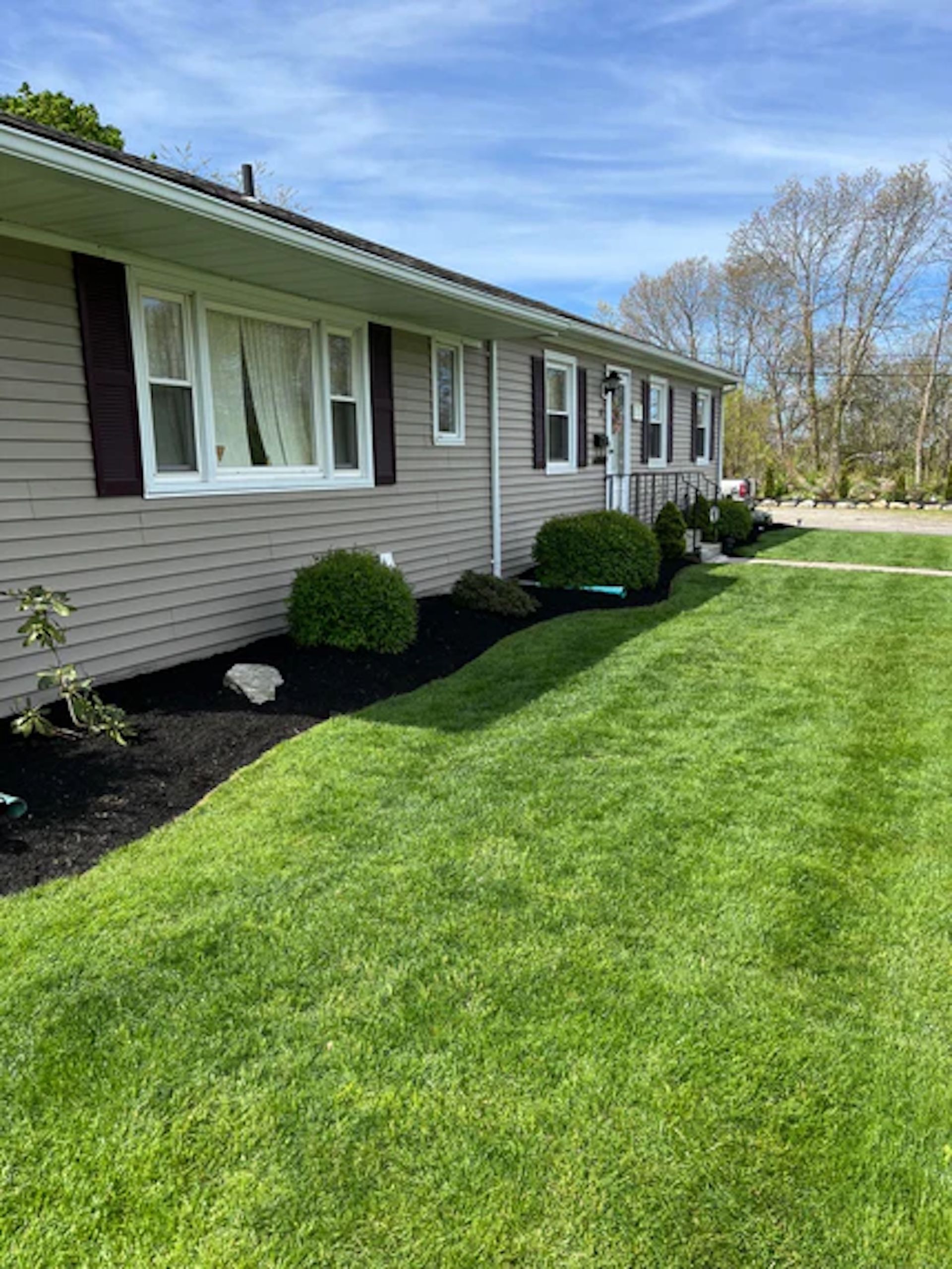 A single-story house with beige siding and dark shutters, featuring fresh black mulch and manicured shrubs on a sunny day.