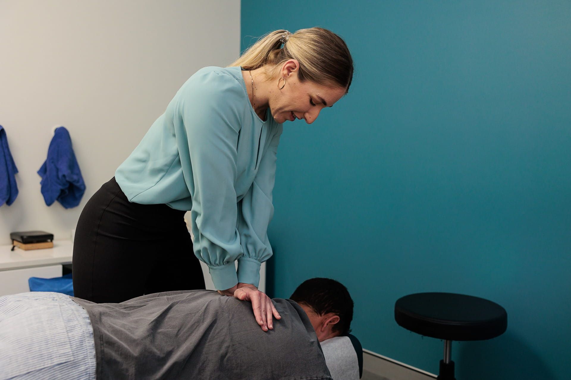Woman in Blue Top Examines a Man Lying Face Down on A Massage Table, in An Office Setting — Grays Chiropractic & Wellness In Marian, QLD
