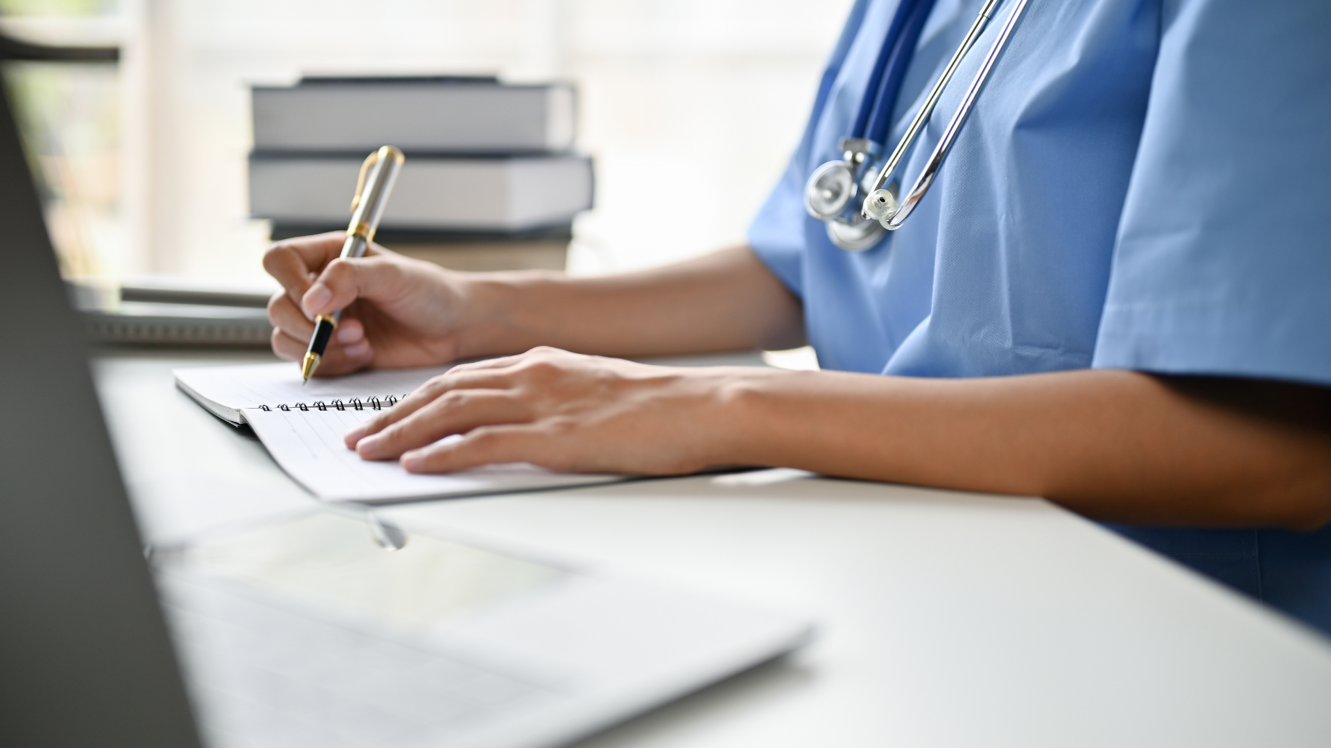 Healthcare worker writing notes at a desk with a stethoscope visible