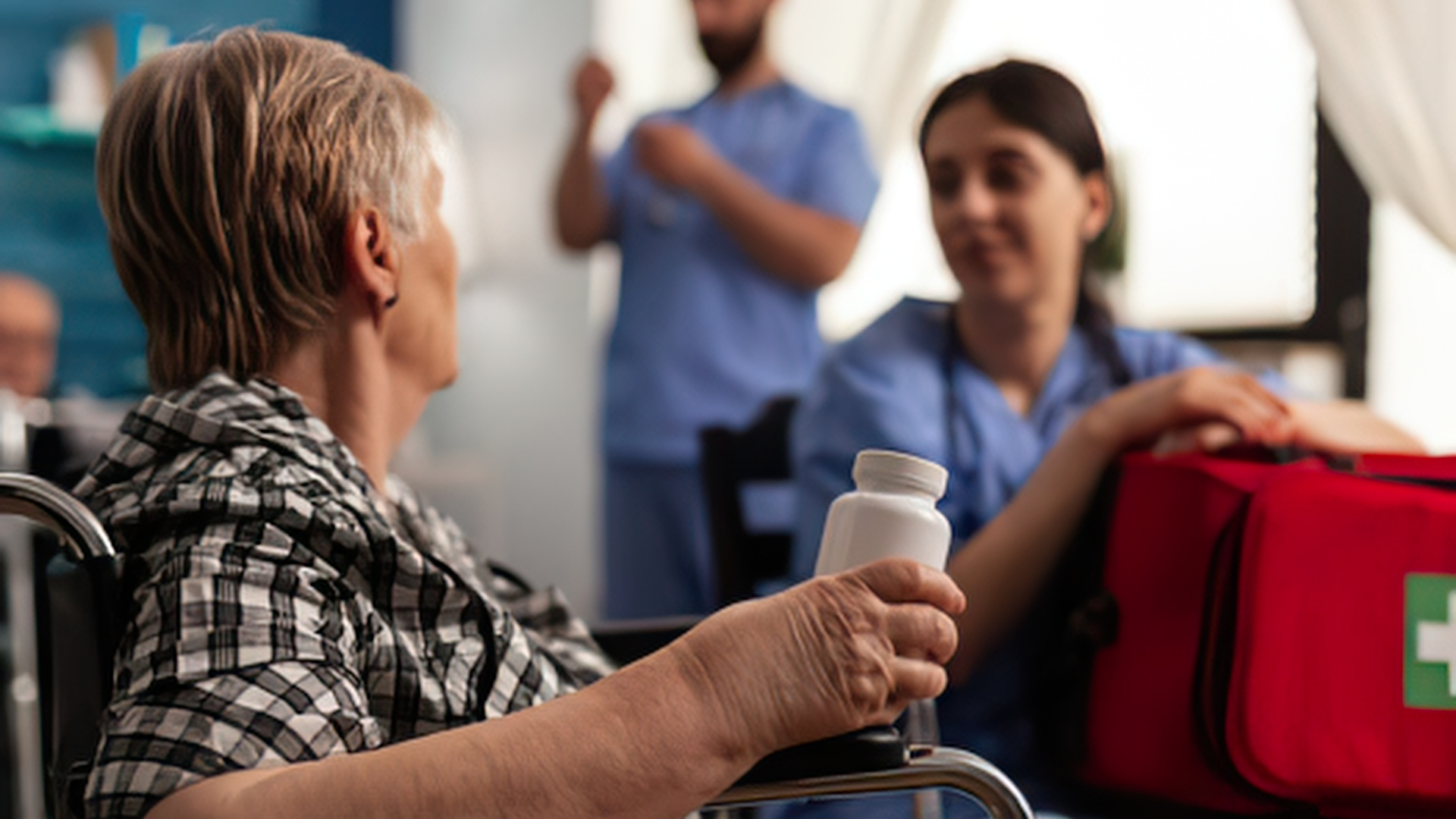 Patient in wheelchair holding a pill bottle, speaking with a nurse beside a red first-aid bag.