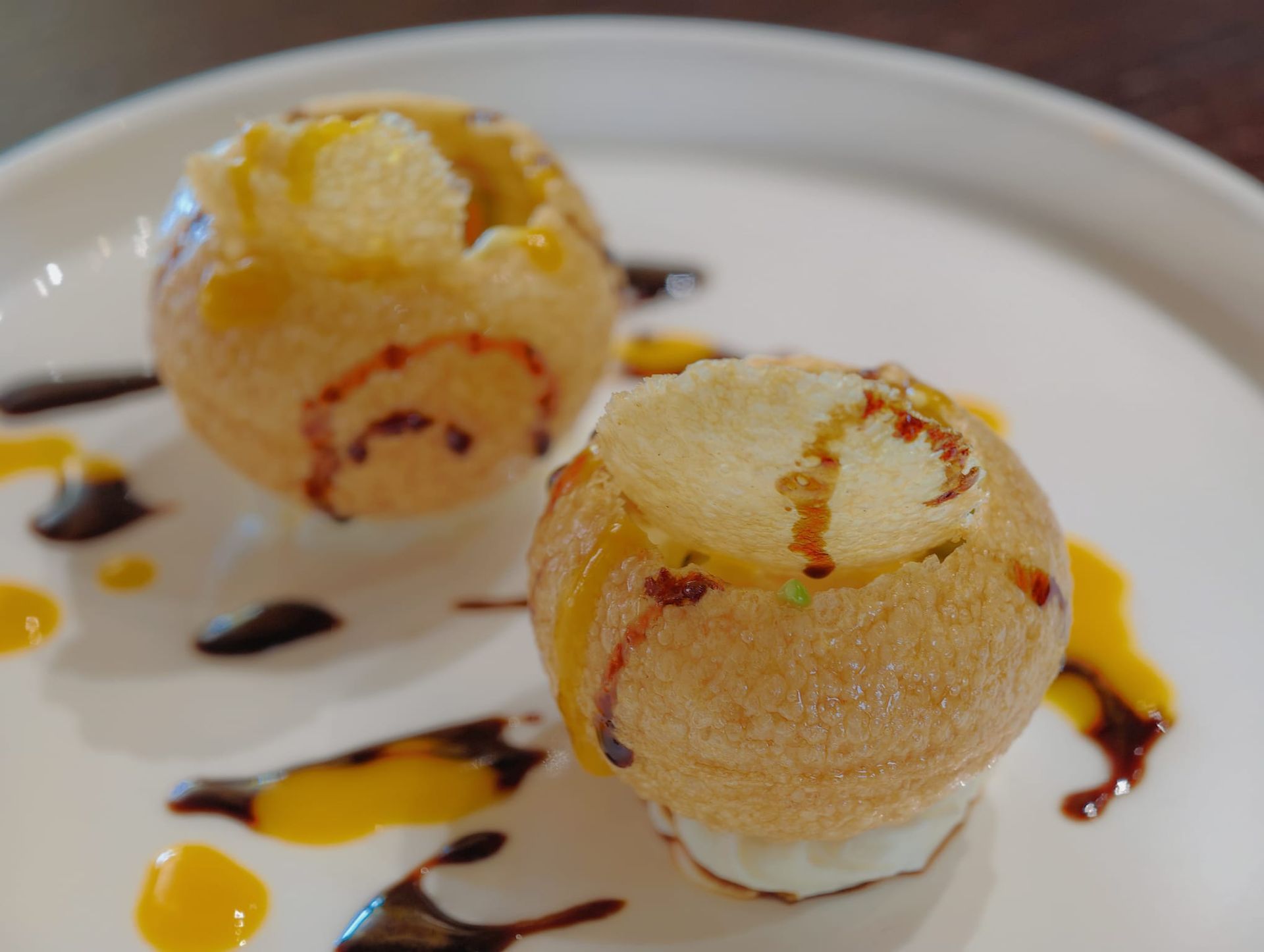 Due sfere di dolce ripiene di crema, guarnite con caramello e cioccolato, su un piatto bianco