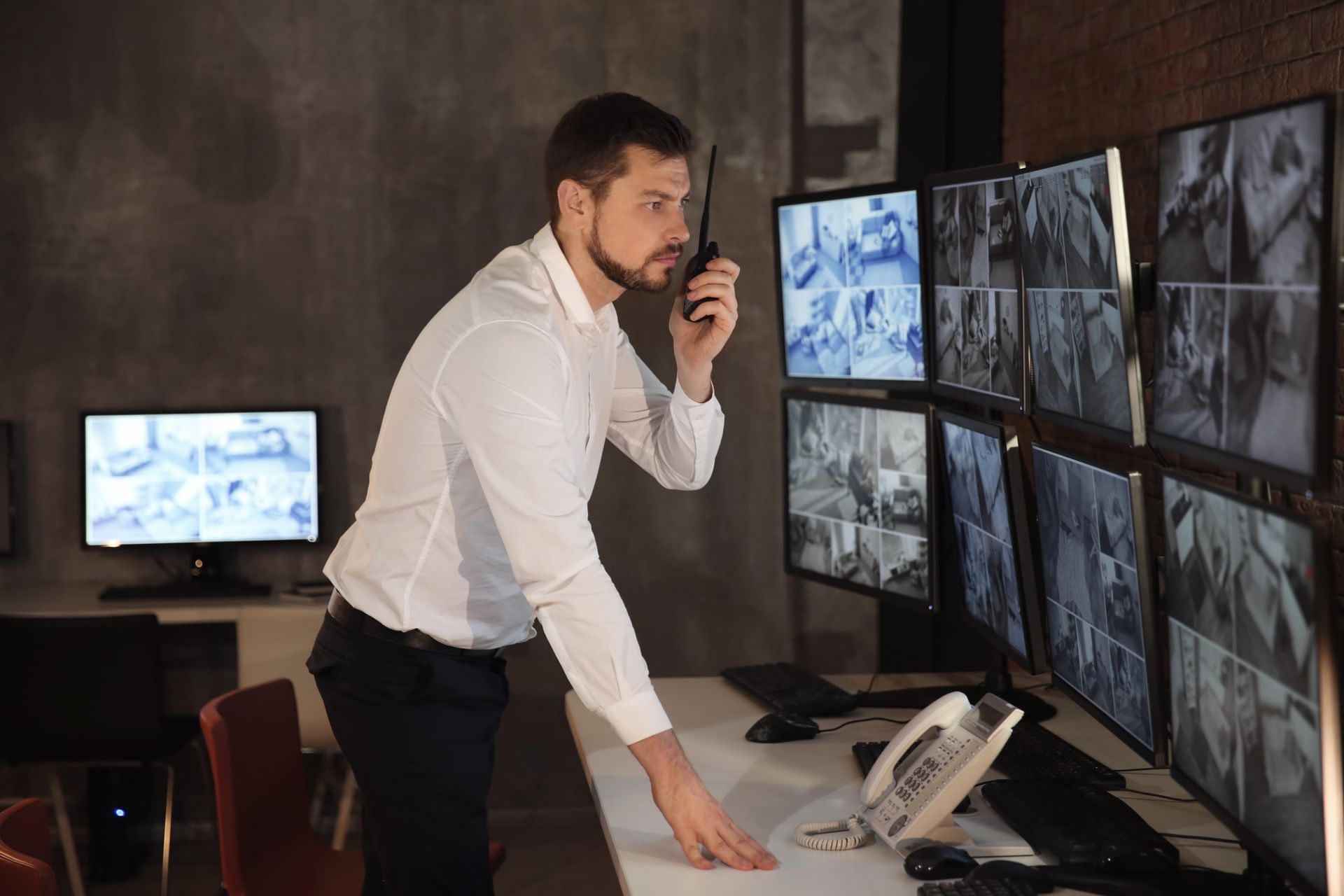 Man in white shirt talking on walkie-talkie, monitoring security cameras. Office setting.