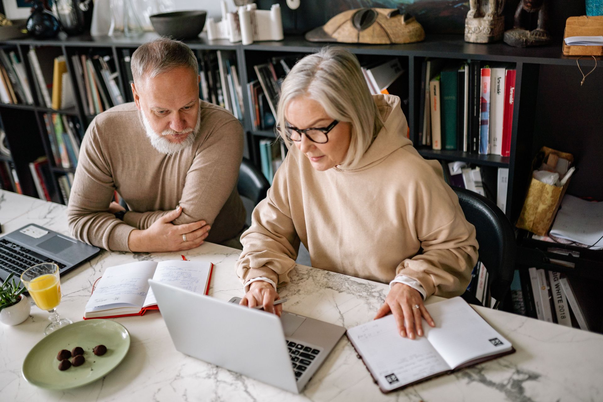 Two people working together at a desk with a laptop, notebooks, and coffee in a home office