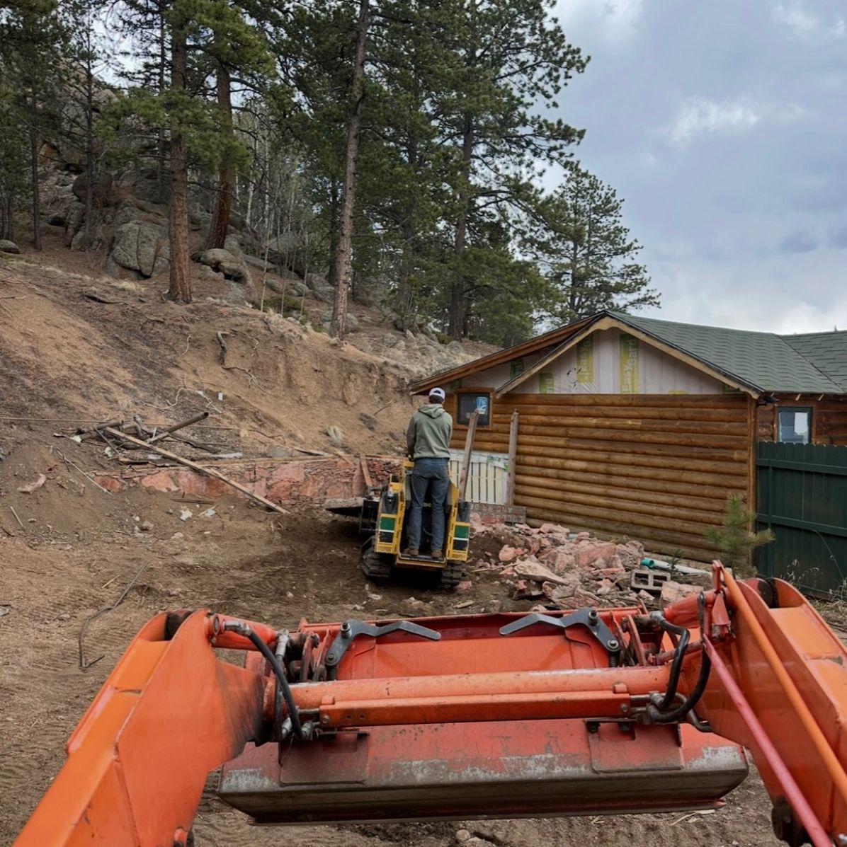 Orange excavator at a wooded hillside near a log cabin, with a worker standing in the bucket.