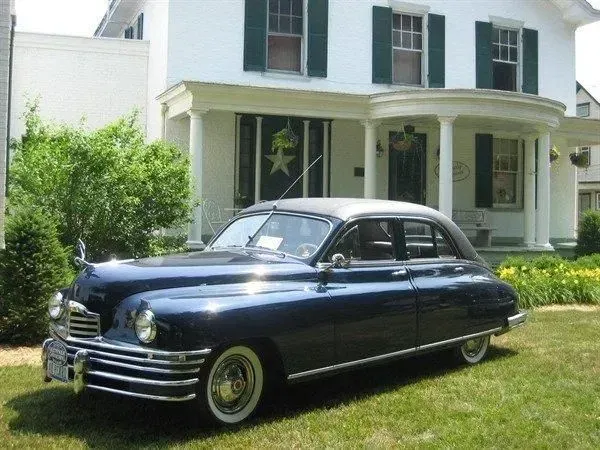 Dark blue vintage Packard car parked on a green lawn in front of a white house with a porch.