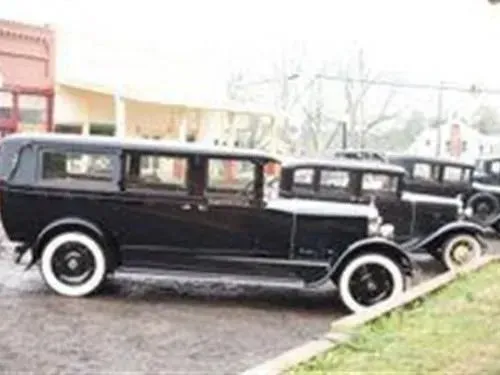 Black vintage cars parked in front of a building; white-wall tires, overcast day.