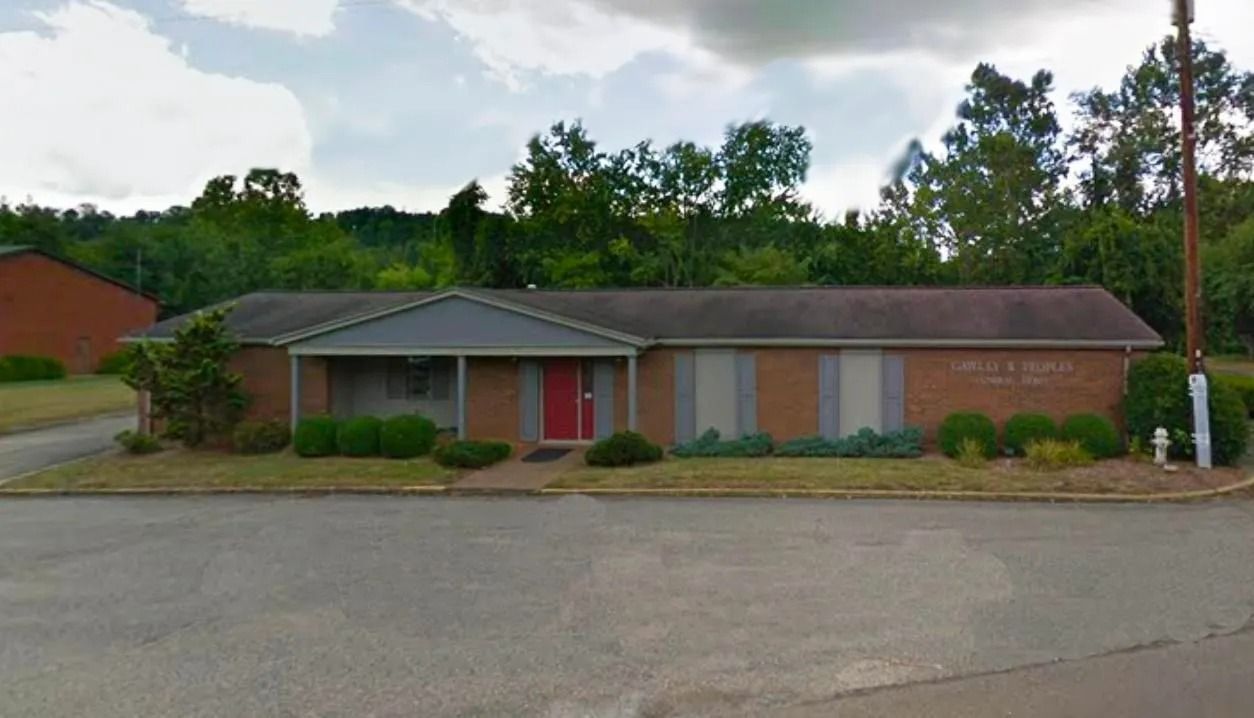 Long, low, brick building with a red door, gray awning, and small windows, surrounded by green grass and trees.
