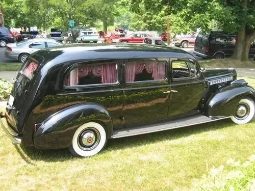 Black vintage hearse with white-walled tires parked on grass, windows with pink curtains, parked near other vehicles.