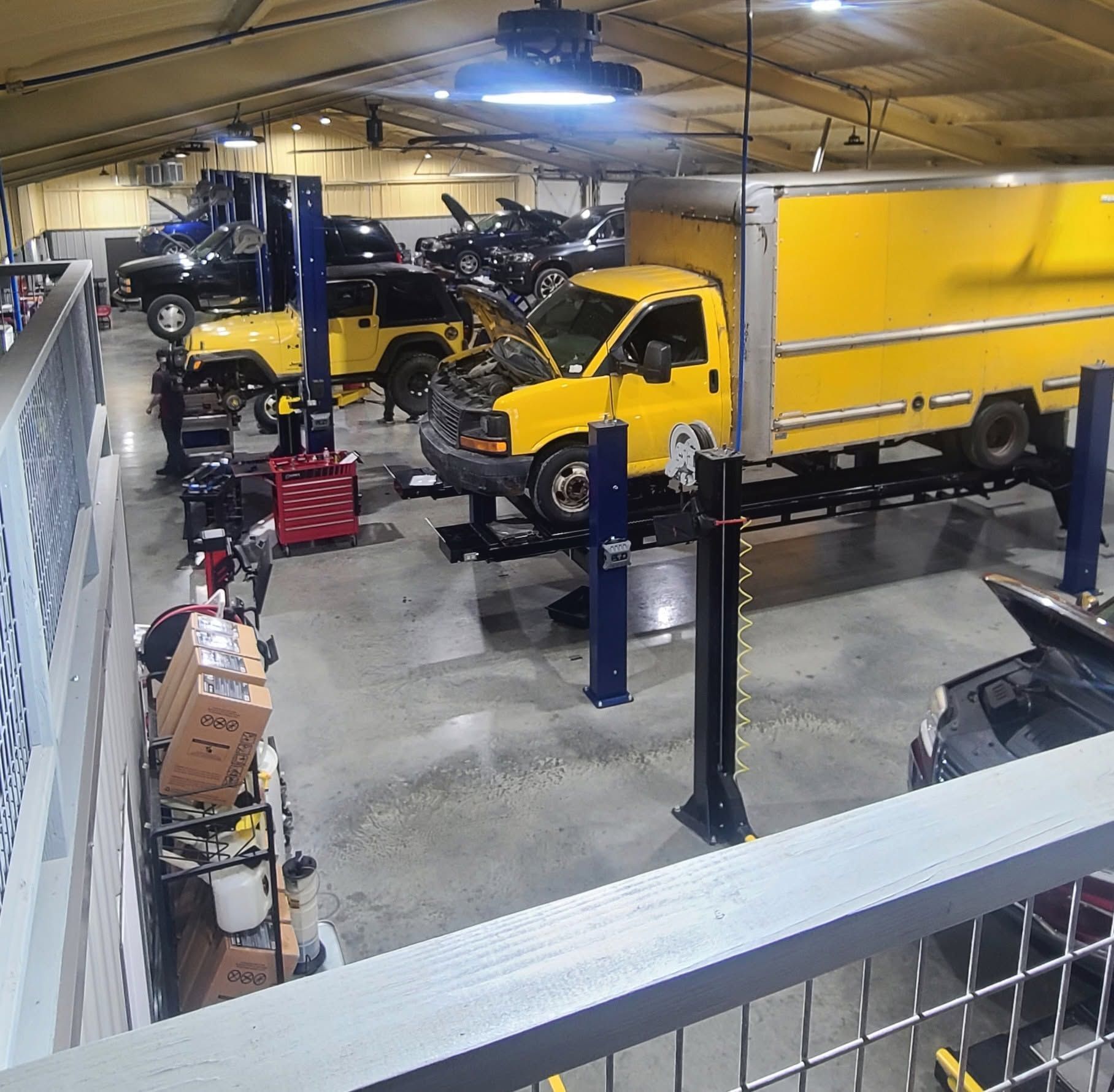 Indoor auto shop with several yellow trucks and cars, some on lifts, under bright overhead lights