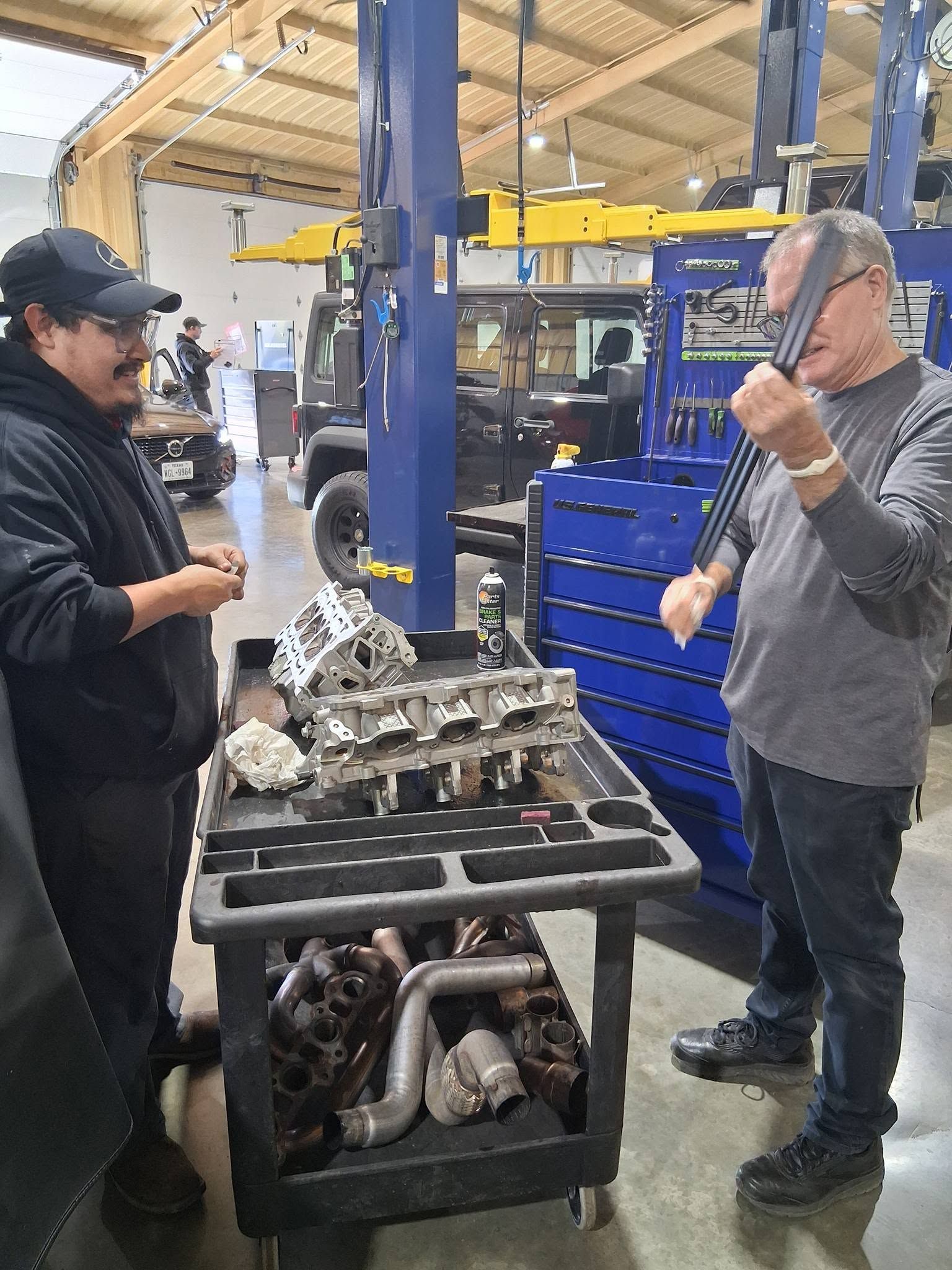 Two workers talking in a factory beside a cart of metal parts and machinery.