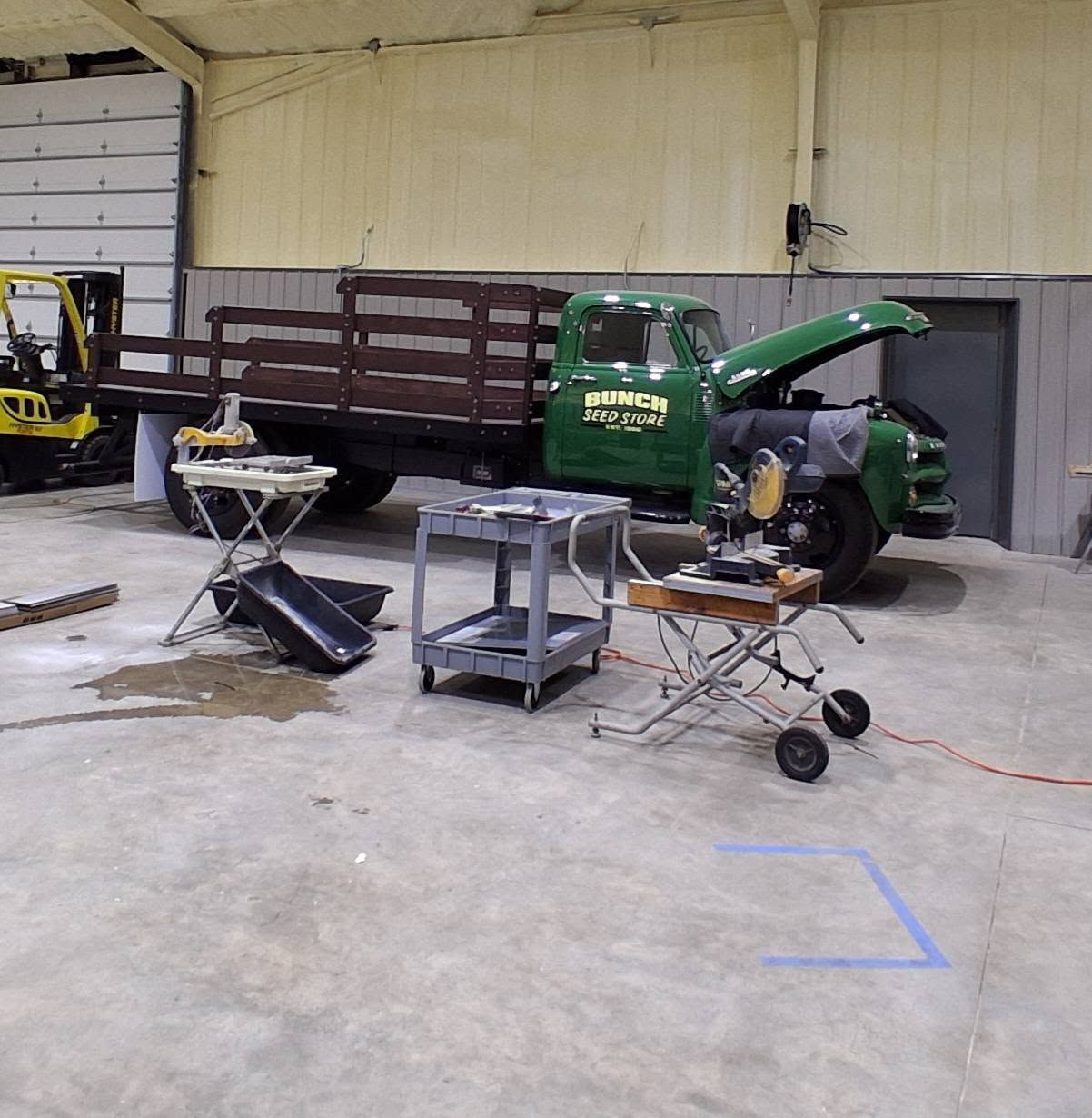 Green utility truck in a warehouse beside a cart, tables, and tools on a concrete floor