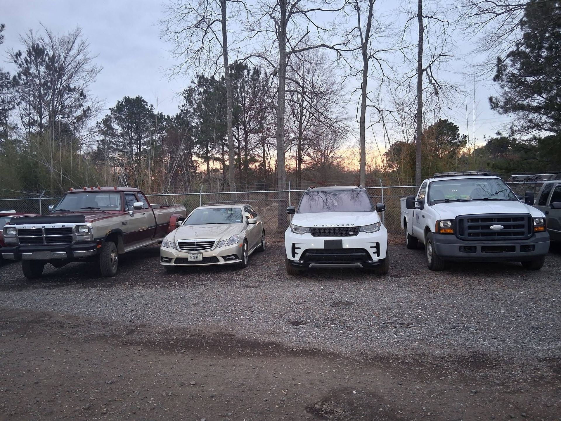 Four parked vehicles on a gravel lot at dusk with bare trees in the background.