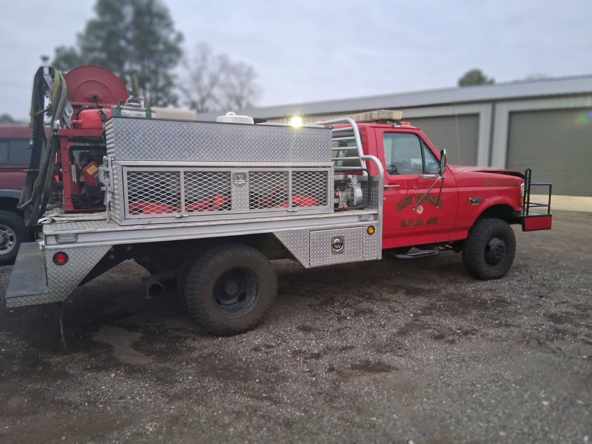 Red utility truck with silver equipment bed parked on gravel beside a building.