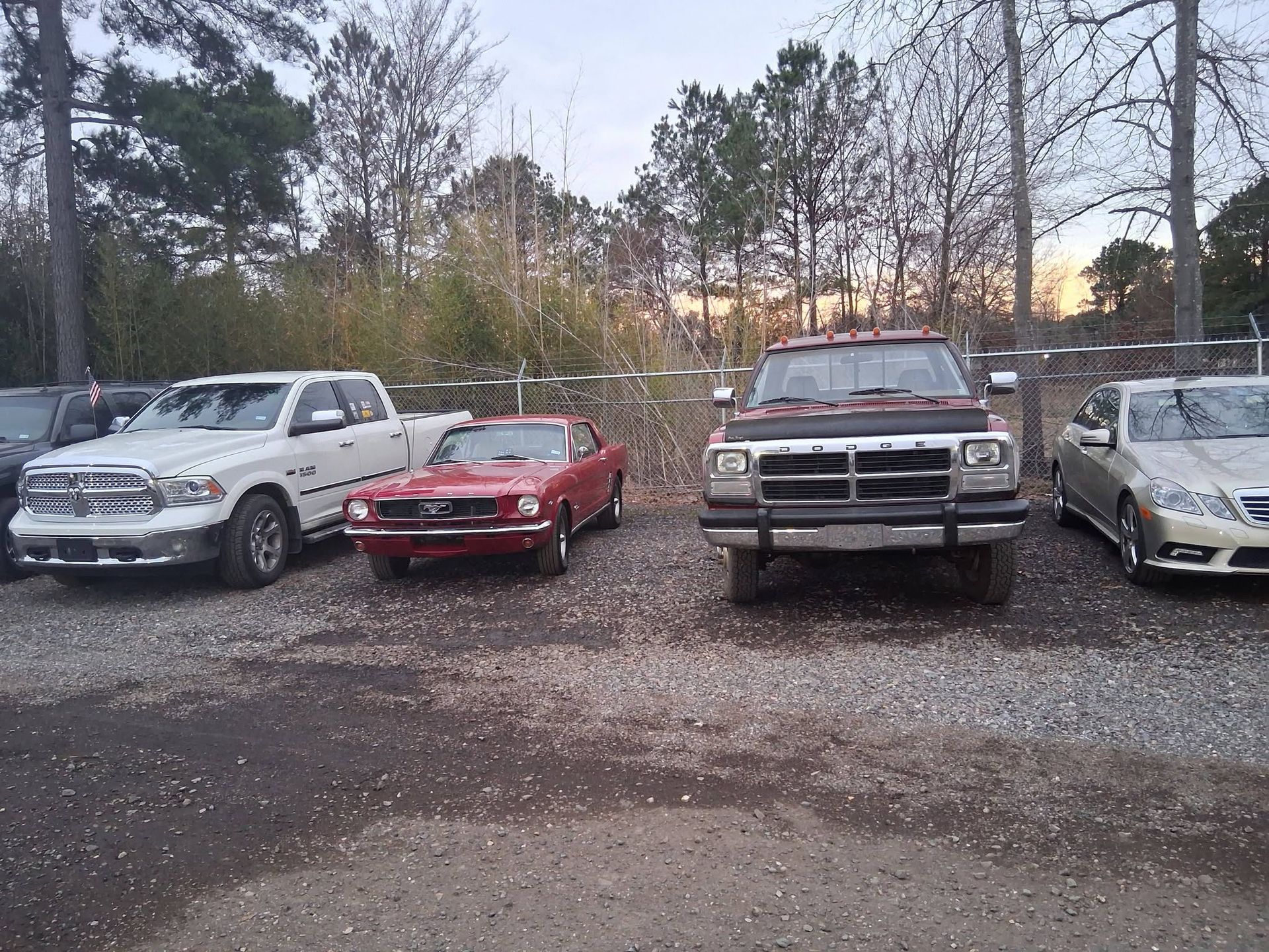 Parked vehicles in a gravel lot with trees in the background, including a red convertible and white trucks.