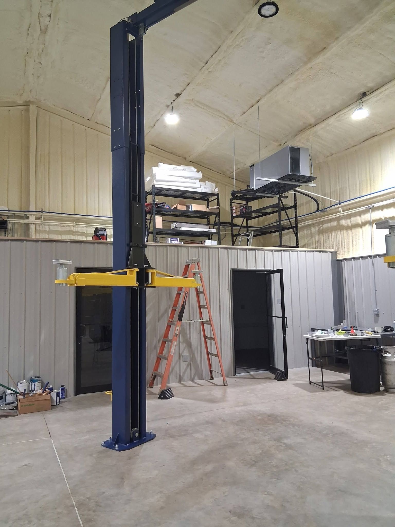 Blue hydraulic lift in a warehouse workshop with a yellow arm and ladder nearby.