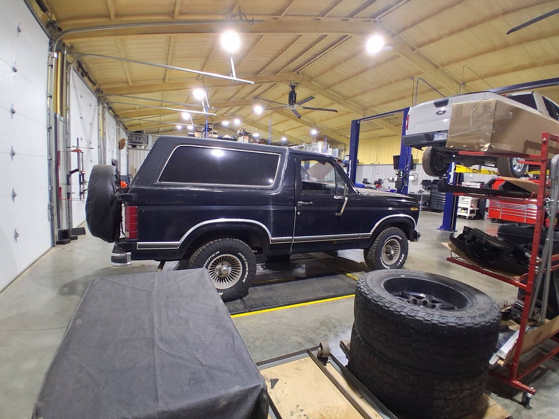 Black SUV in an auto repair shop with tires and tools under bright overhead lights.