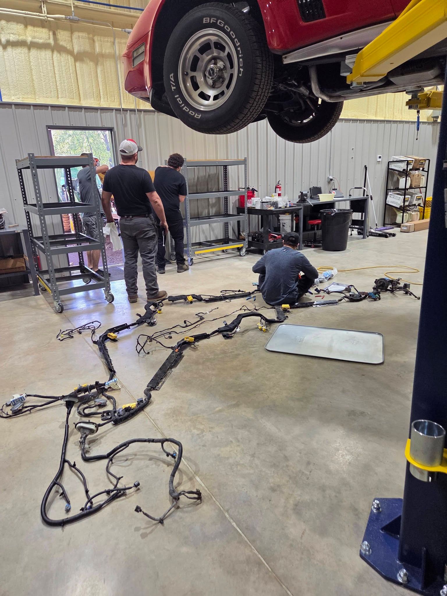 Auto shop with car lifted overhead and three mechanics working on tools and wiring on the floor