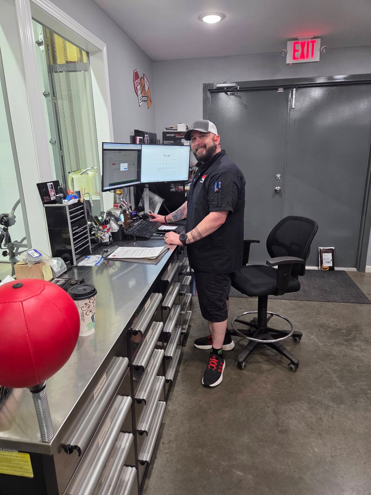 Person at a workbench in a garage, standing by tools and a computer, with a red exercise ball nearby.