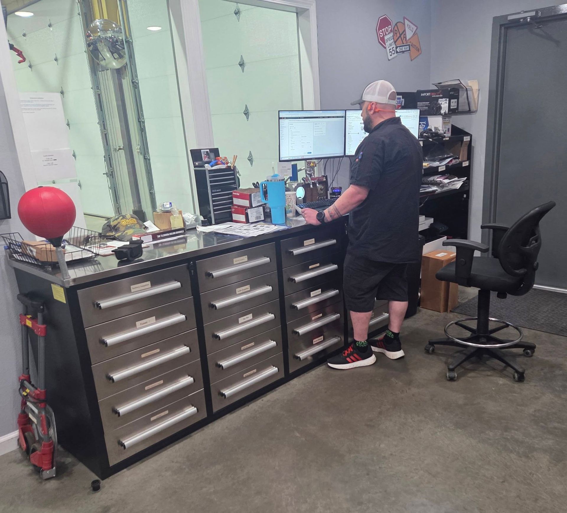 Mechanic in a workshop standing at a workbench with computer monitors and drawers, surrounded by tools and equipment