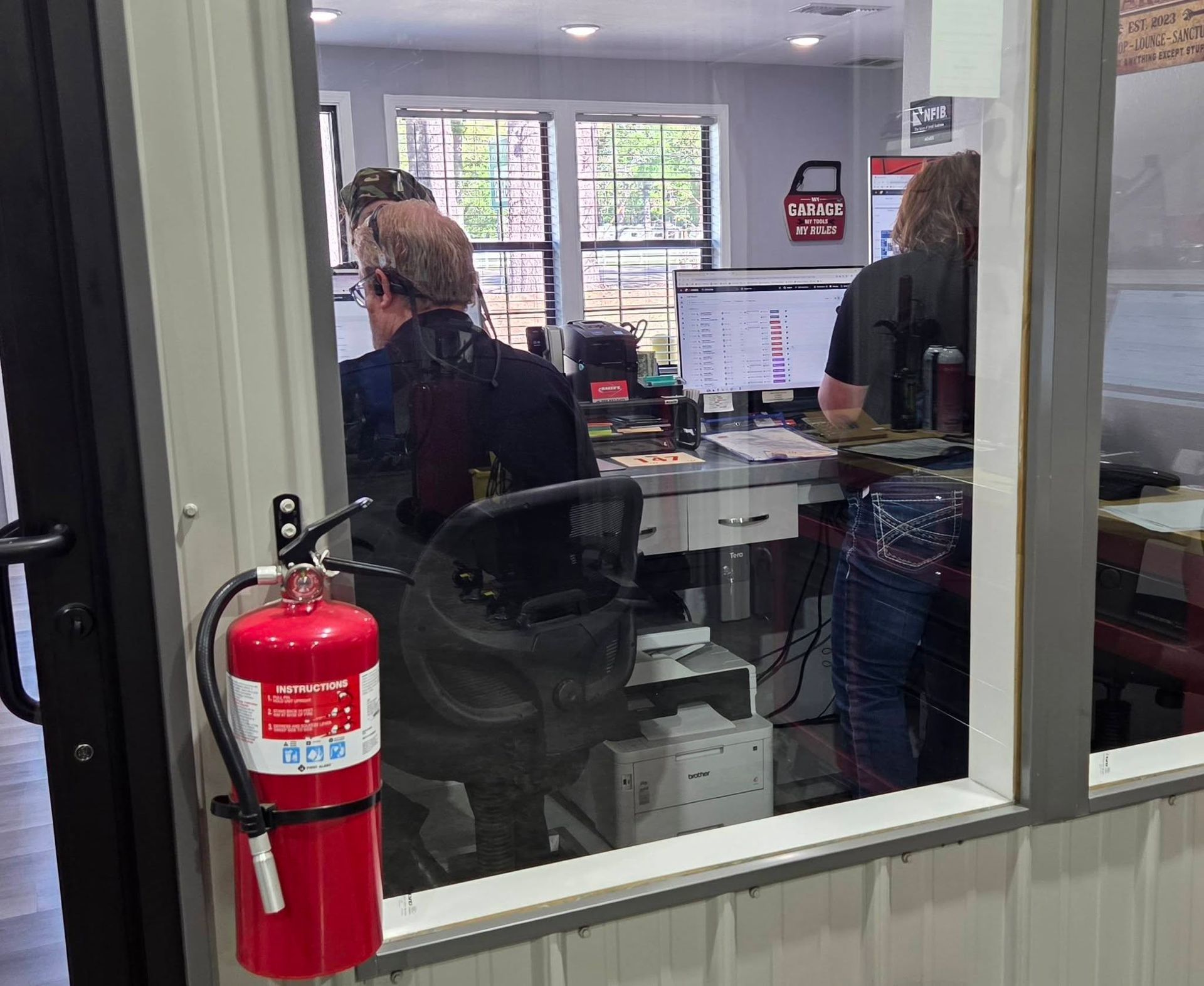 Office scene viewed through a window, with people at desks, monitors, and a red fire extinguisher in the foreground