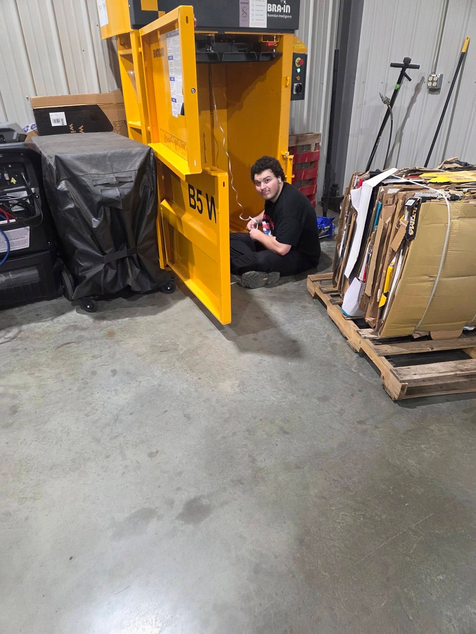 Person crouches beside a yellow industrial machine in a warehouse, with stacked pallets and cardboard boxes nearby.