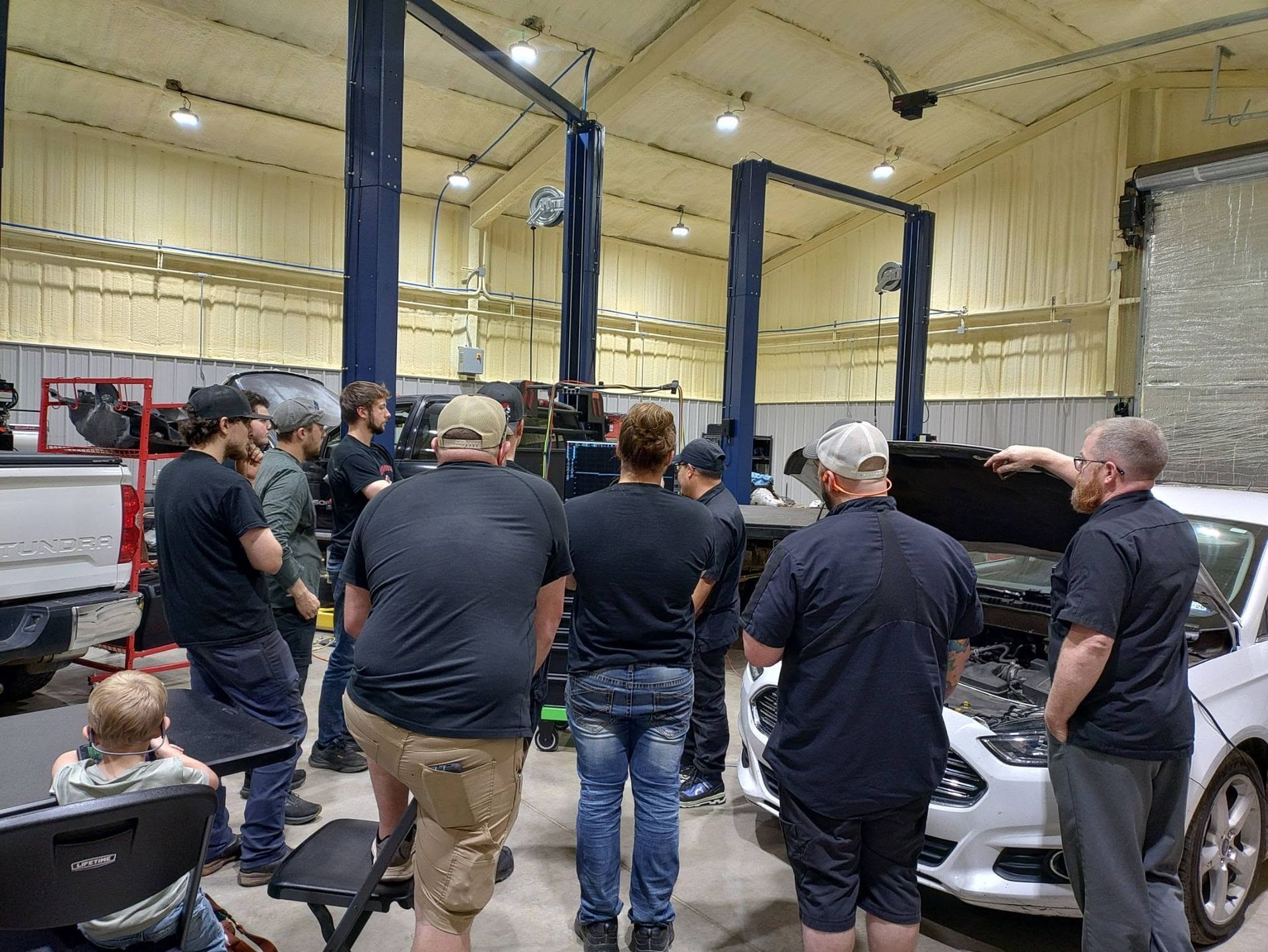 Group of people gathered around a silver car in a workshop with lifts and equipment.