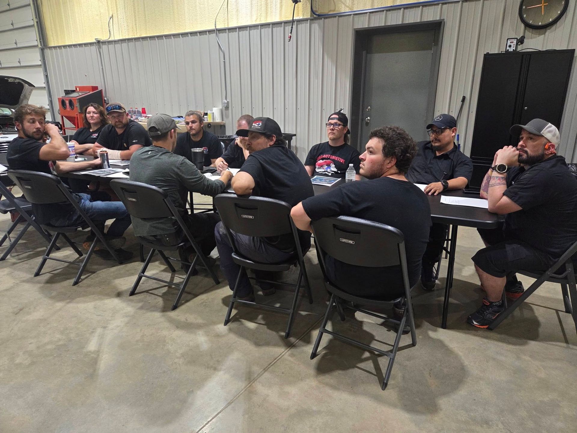 People seated at long tables in a warehouse-style room, talking over a meal or meeting.