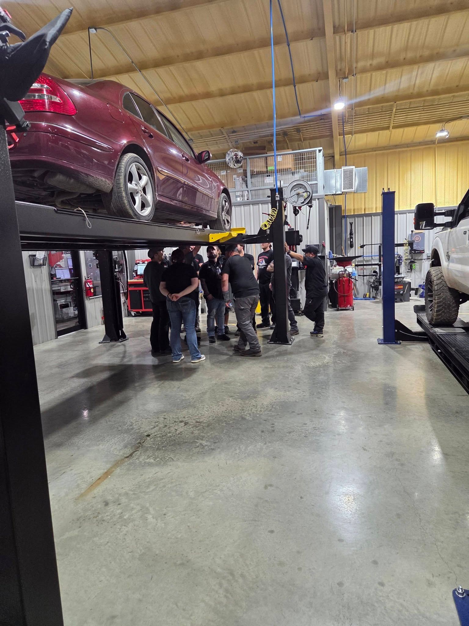 Red car on a lift in an auto shop with several people gathered underneath.