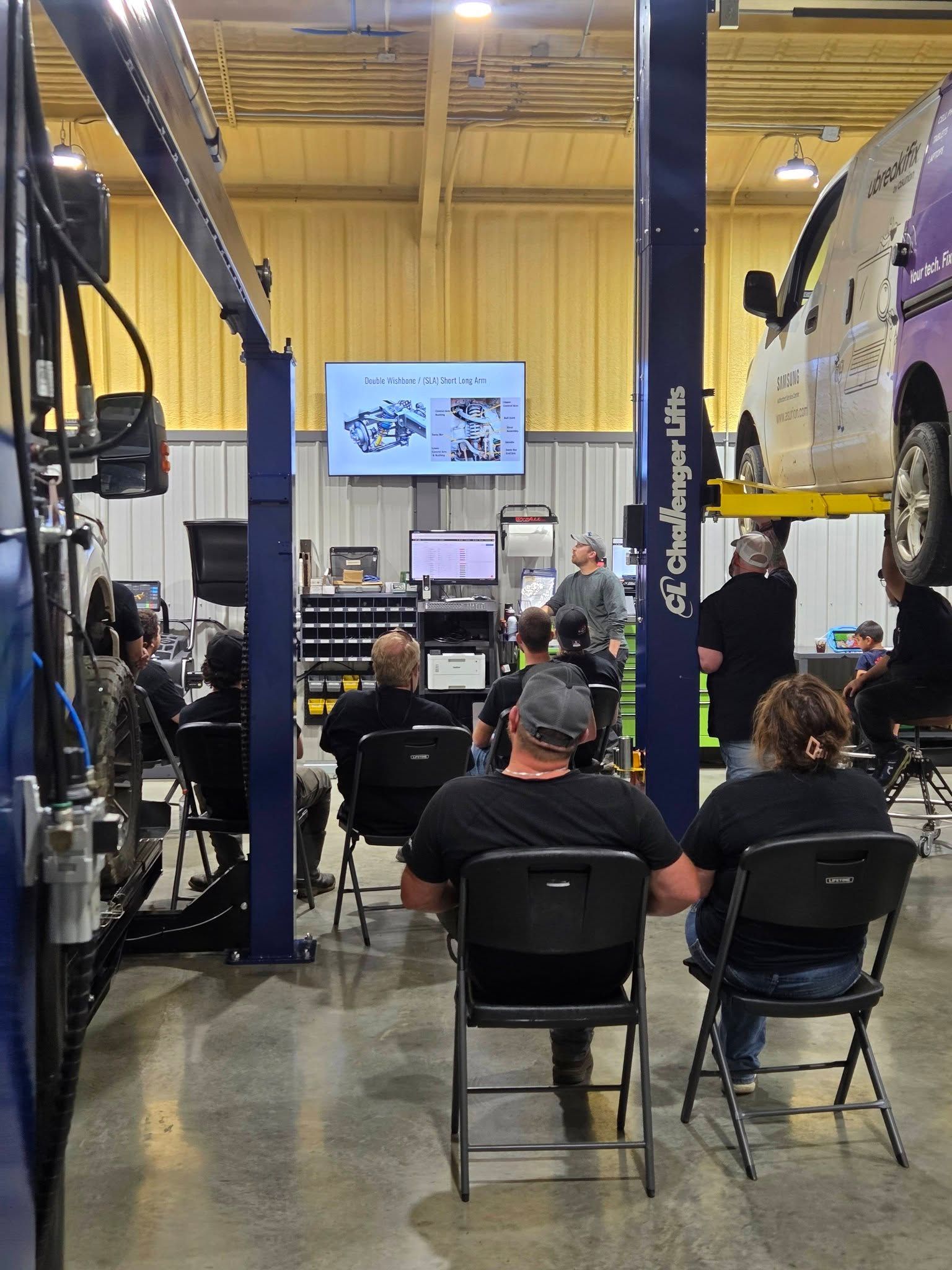 Workshop audience seated before a presentation screen in an auto garage, with a vehicle on a lift.
