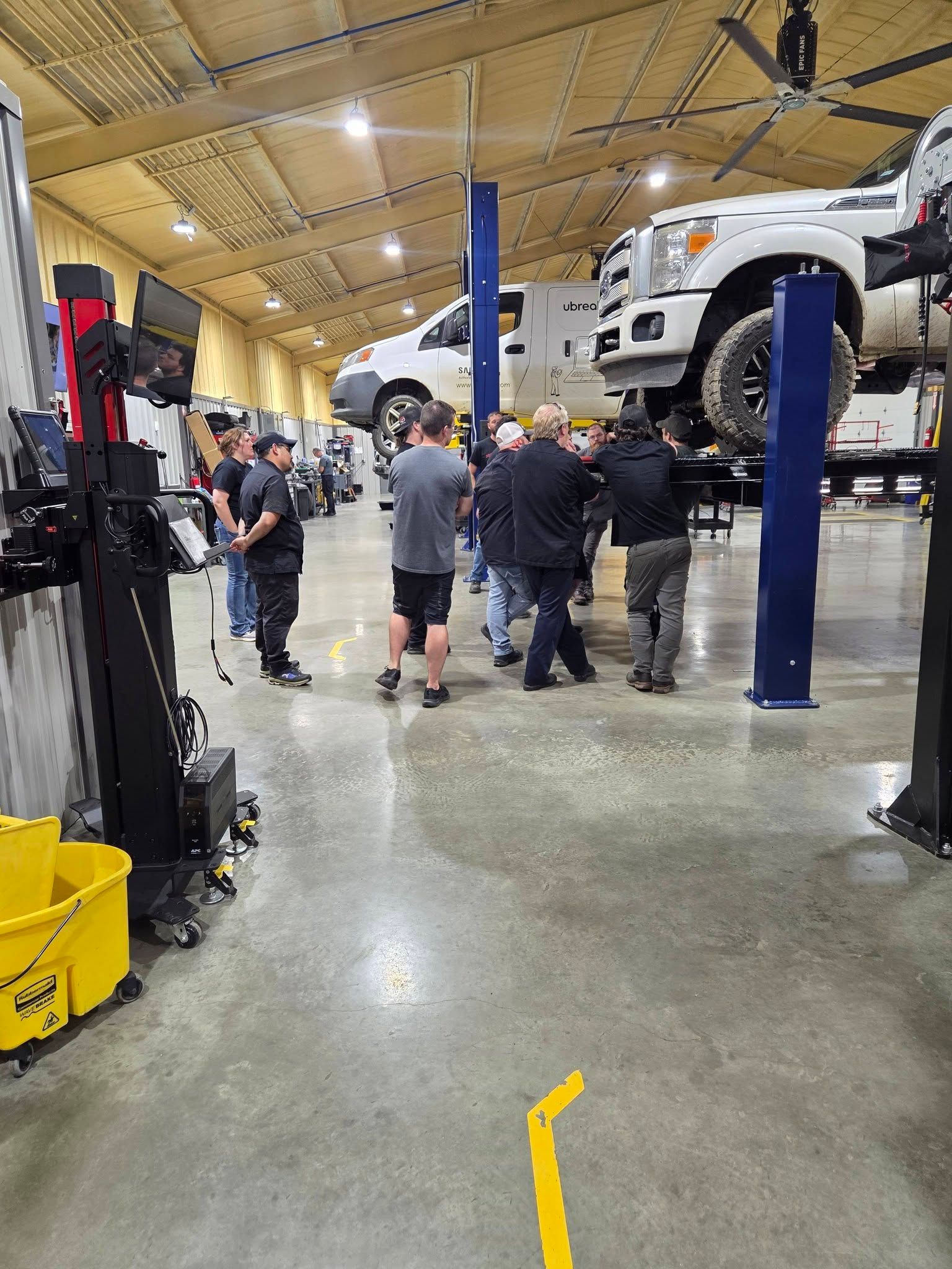 People inspecting lifted white vehicles in an auto repair garage with blue posts and yellow lights.