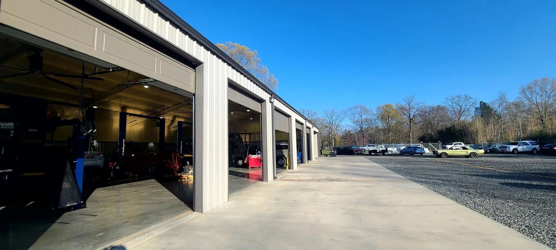 Row of open garage bays beside a paved lot, with cars and trees under a bright blue sky