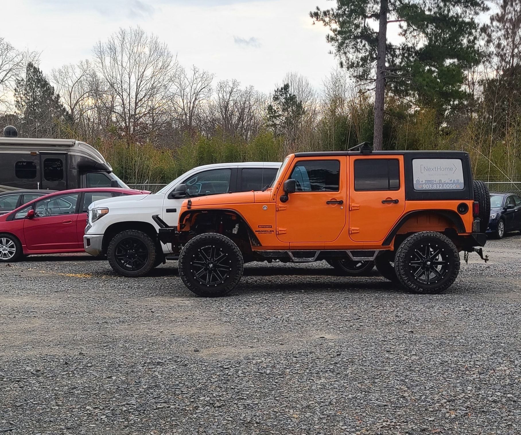 Orange lifted Jeep parked beside a red car on a gravel lot with trees in the background