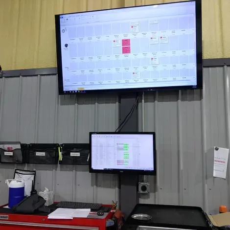 Workshop wall with two mounted monitors showing schedules above a cluttered desk and tools