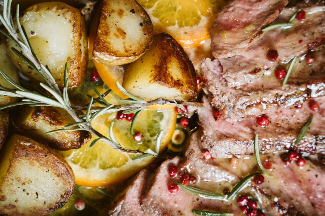 Roasted potatoes, sliced steak, orange slices, and rosemary in a tray, glistening with oil and pink peppercorns.