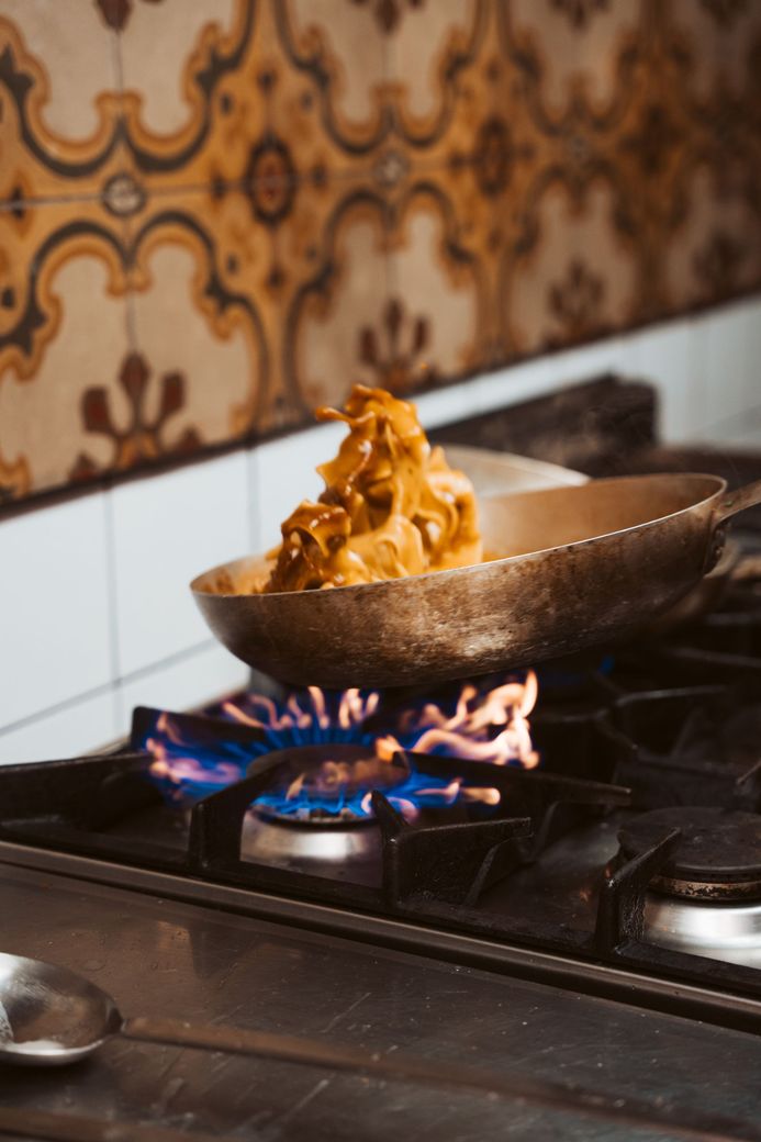 Food cooking in a pan on a lit gas stove, flames visible. Kitchen setting, tiled backsplash.