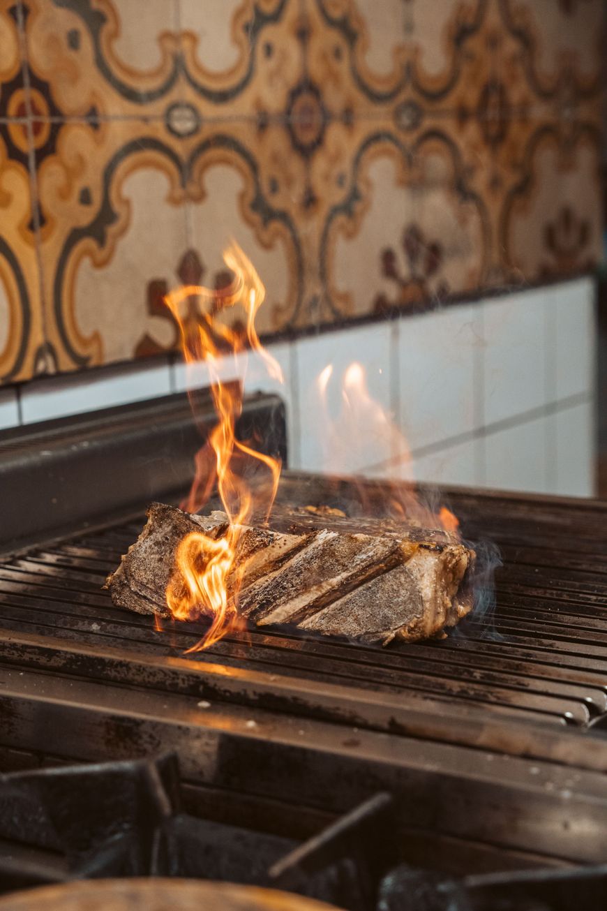 Flames erupting from a large steak grilling on a metal grate; patterned tile background.