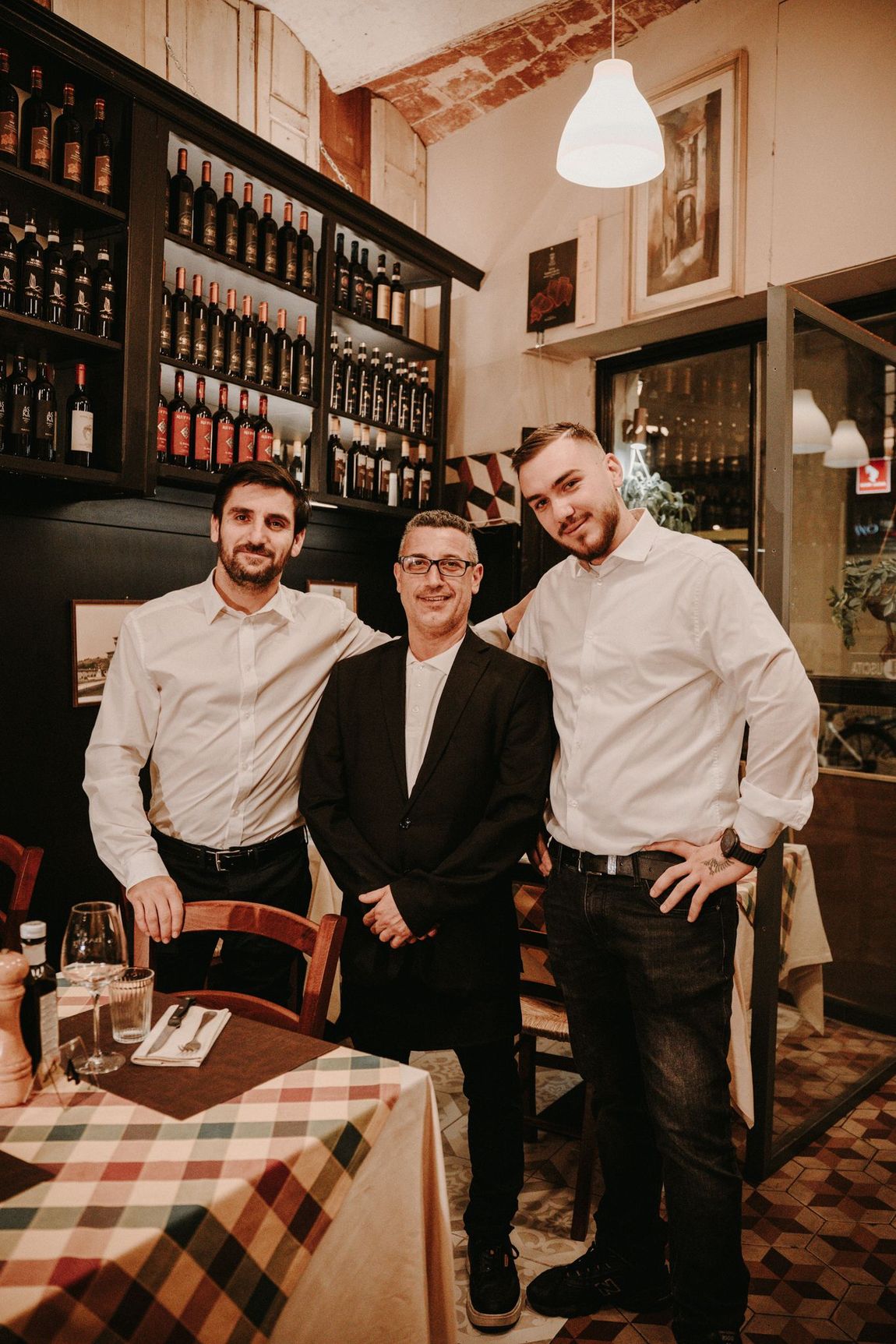 Tre uomini in un ristorante. Due in camicia bianca, uno in abito, in piedi accanto a una tovaglia a quadri. 