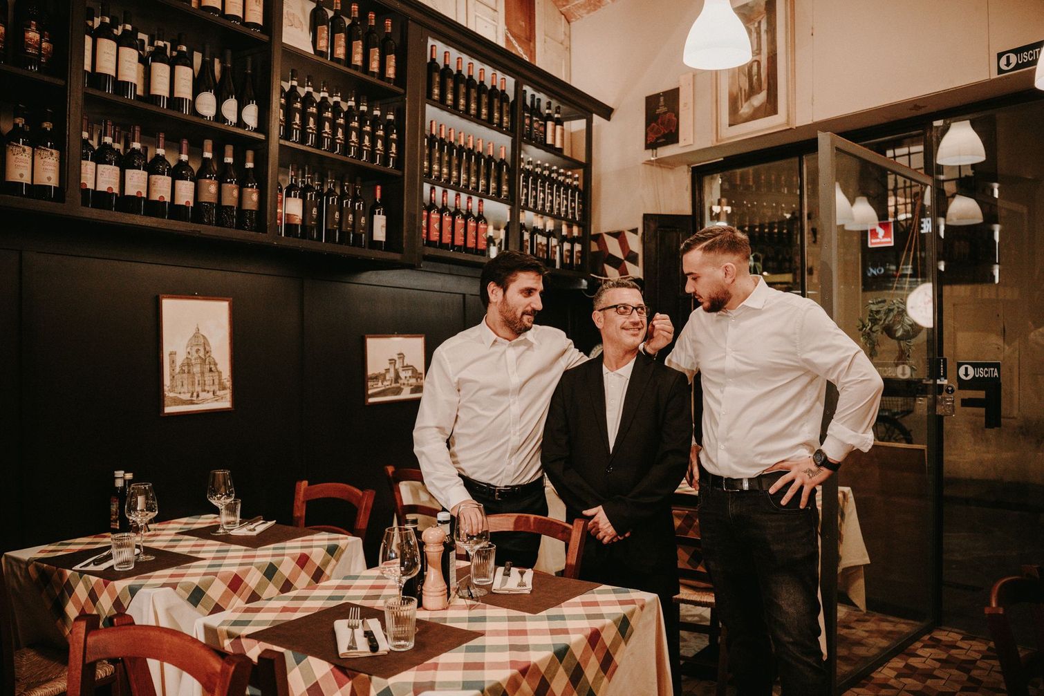 Tre uomini sono in piedi in un ristorante italiano. Uno in giacca e cravatta, due in camicia bianca.